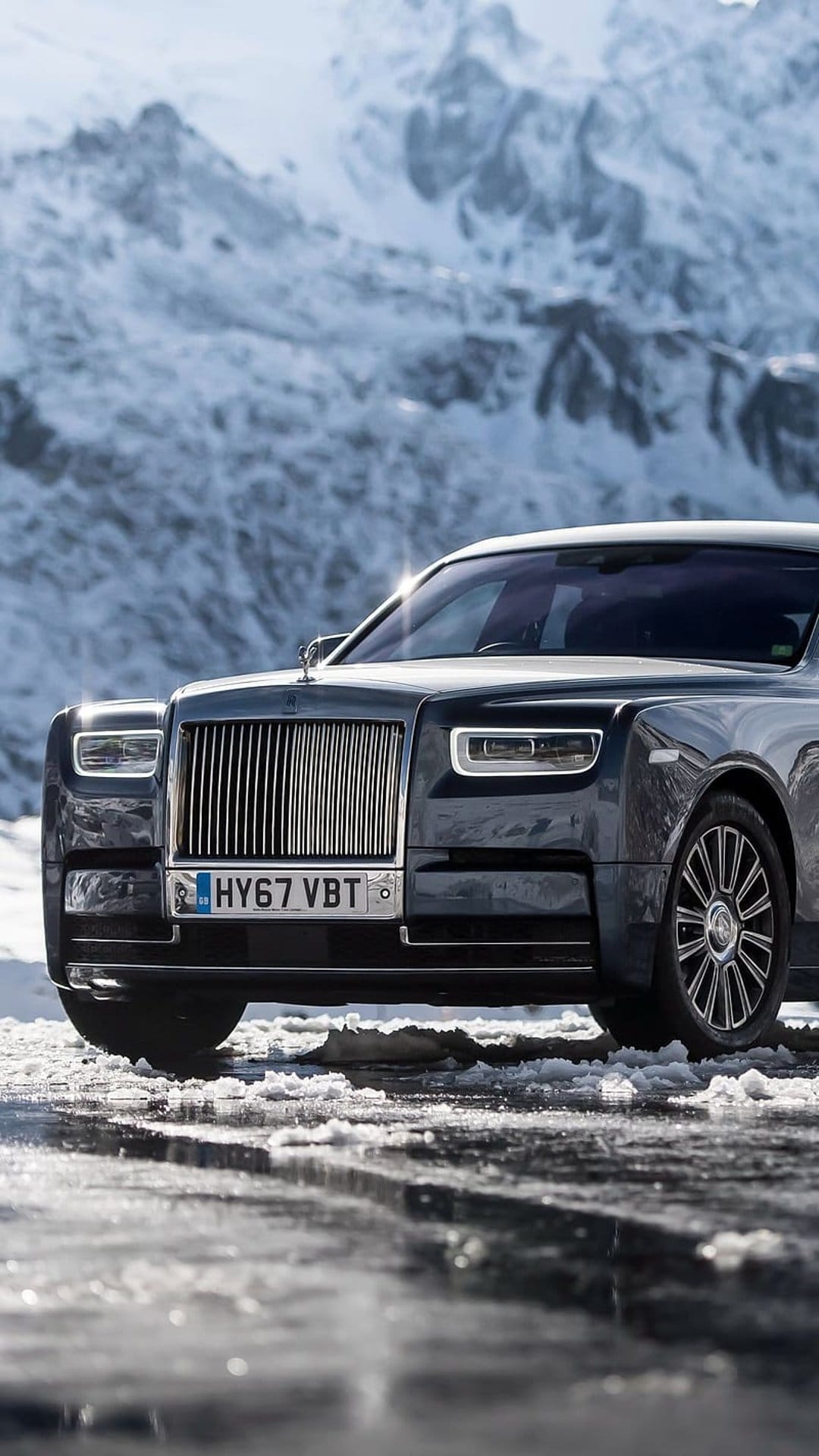 Charcoal Rolls-Royce Phantom parked on a snowy road with sunlit headlights and jagged mountain peaks behind it.