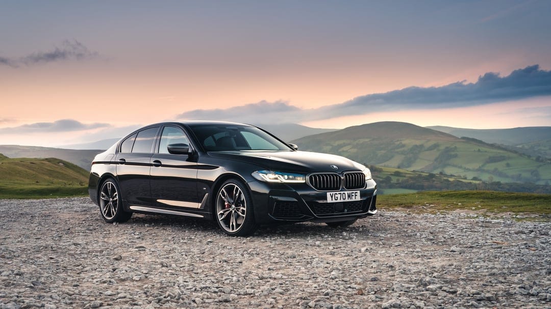Black BMW sedan parked on a gravel hilltop with illuminated headlights against rolling green hills at sunset.