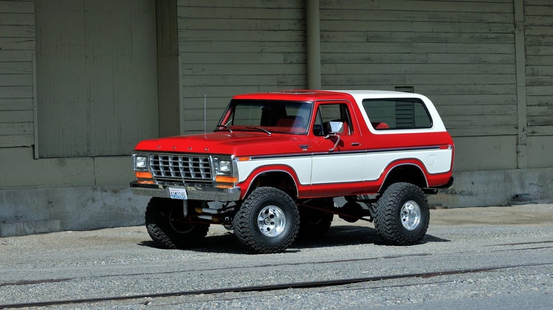 Red and white classic Ford Bronco with a lift kit and large tires parked against a weathered wooden wall.