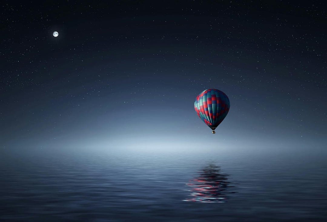 Hot air balloon drifting over calm water at night with a bright moon and stars reflecting on the glassy surface.