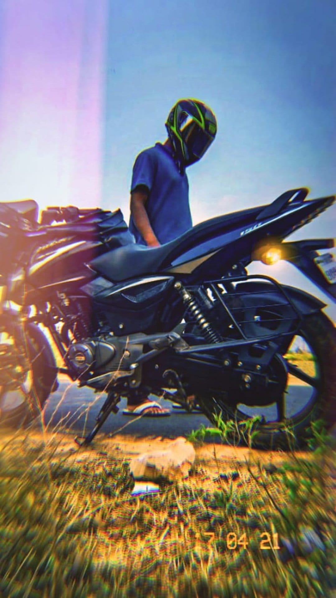 Man in a motorcycle helmet stands by a black bike on a grassy roadside under a clear sky with a retro date stamp.
