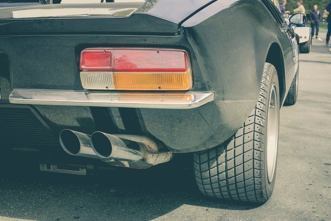 Classic sports car rear with polished chrome exhaust, red tail lights, and diamond tread tires at a car show.