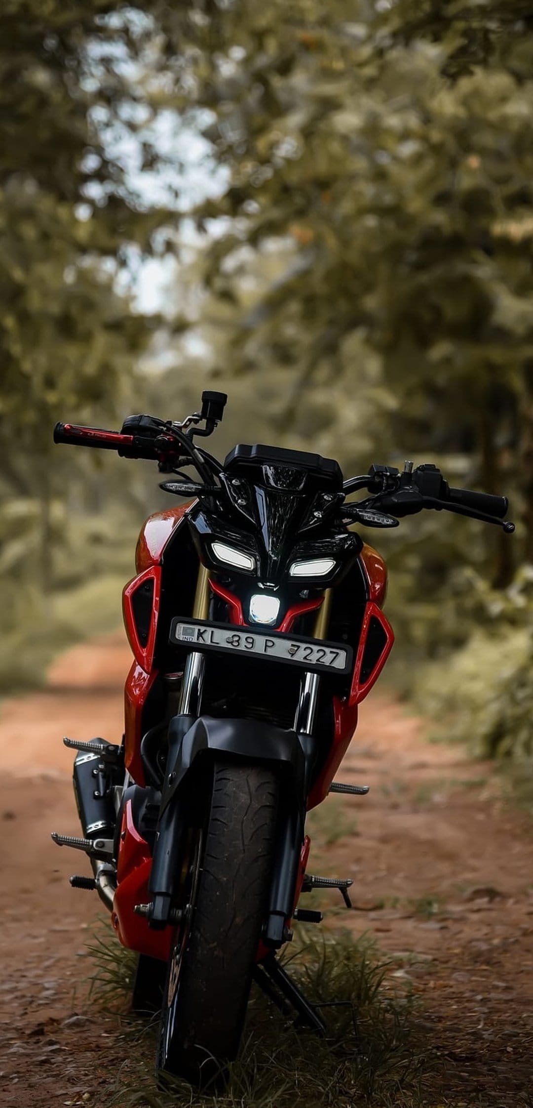 Red Yamaha MT-15 motorcycle with lit LED headlights parked on a narrow dirt path through a lush green forest.