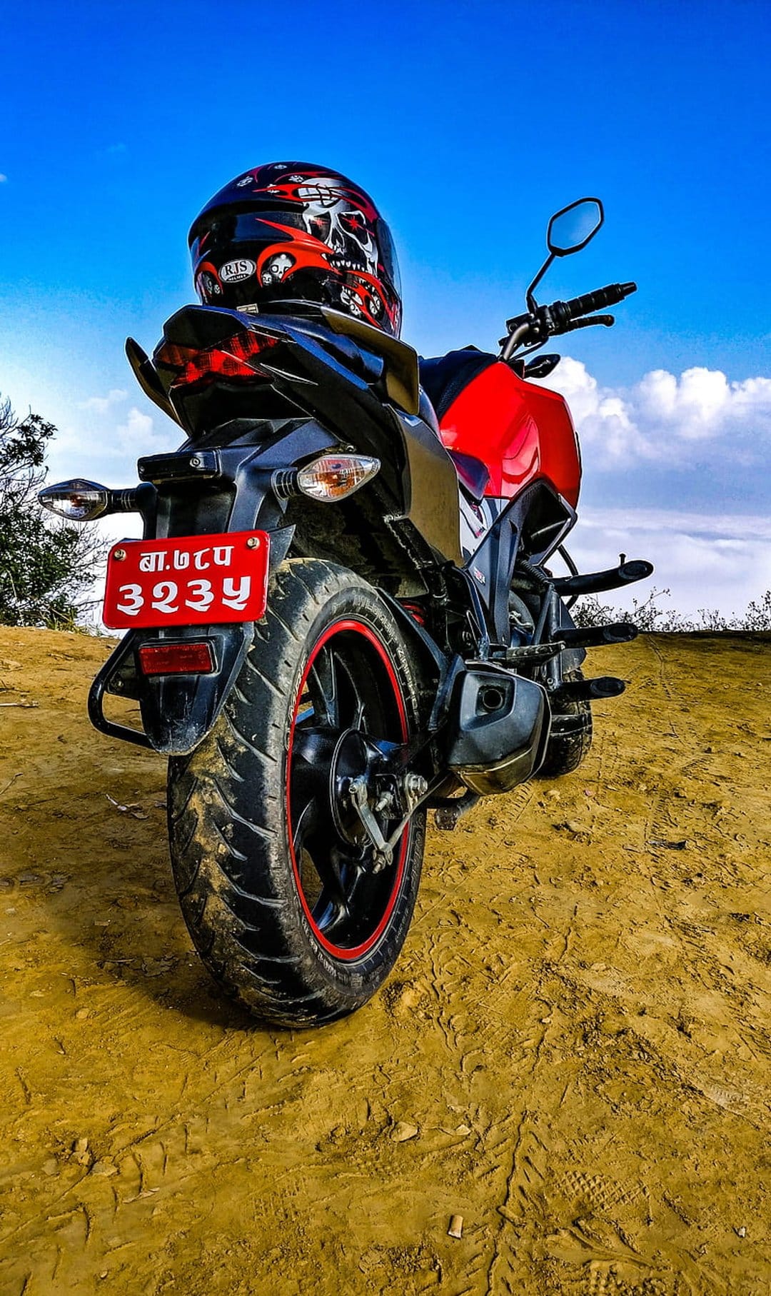 Red motorcycle with a skull helmet parked on a dusty road under a bright blue sky with white clouds.