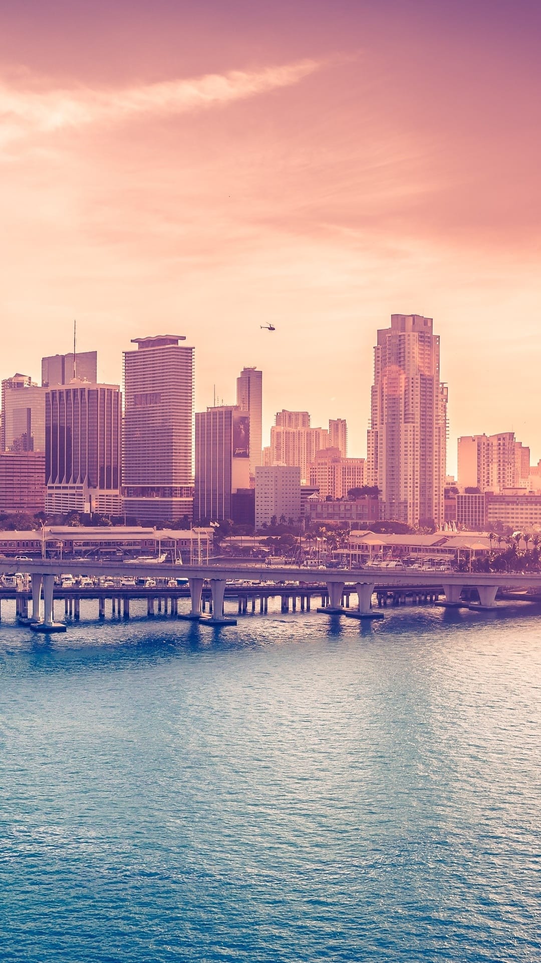 Modern skyscrapers and a multi-level bridge overlook a calm river during a soft pastel sunset with a helicopter.