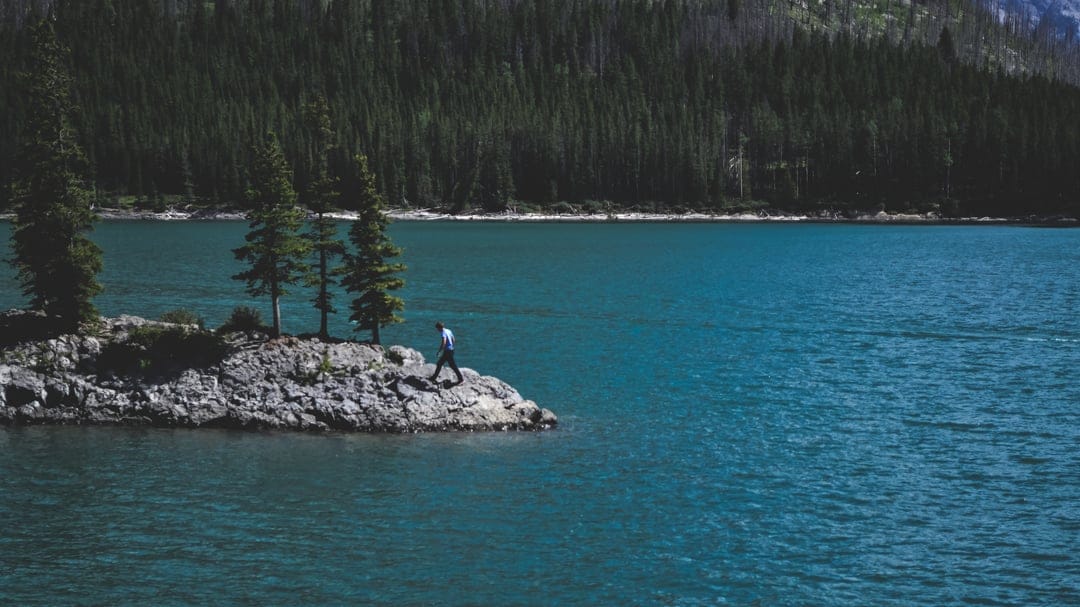 A man walks across a small rocky island with evergreen trees surrounded by vast, rippling turquoise lake water.