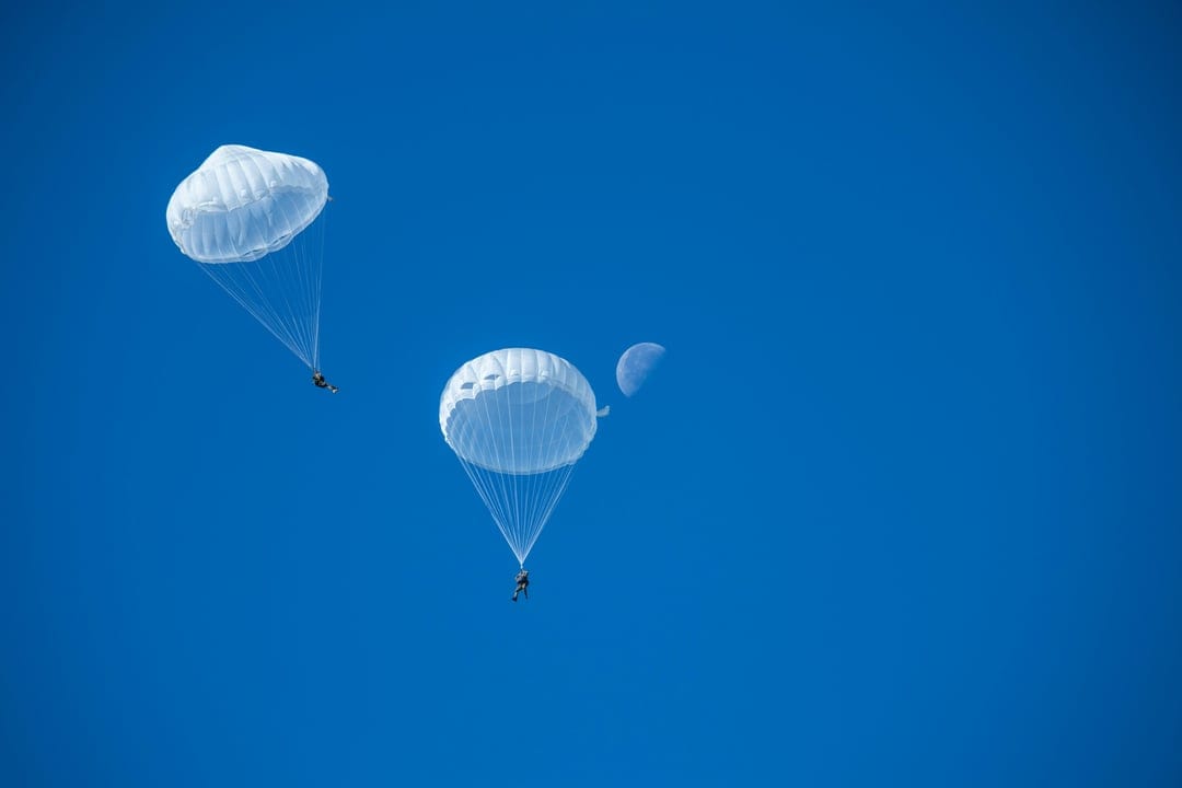 Two white parachutes drift through a vast, cloudless blue sky with small figures hanging from the harness.
