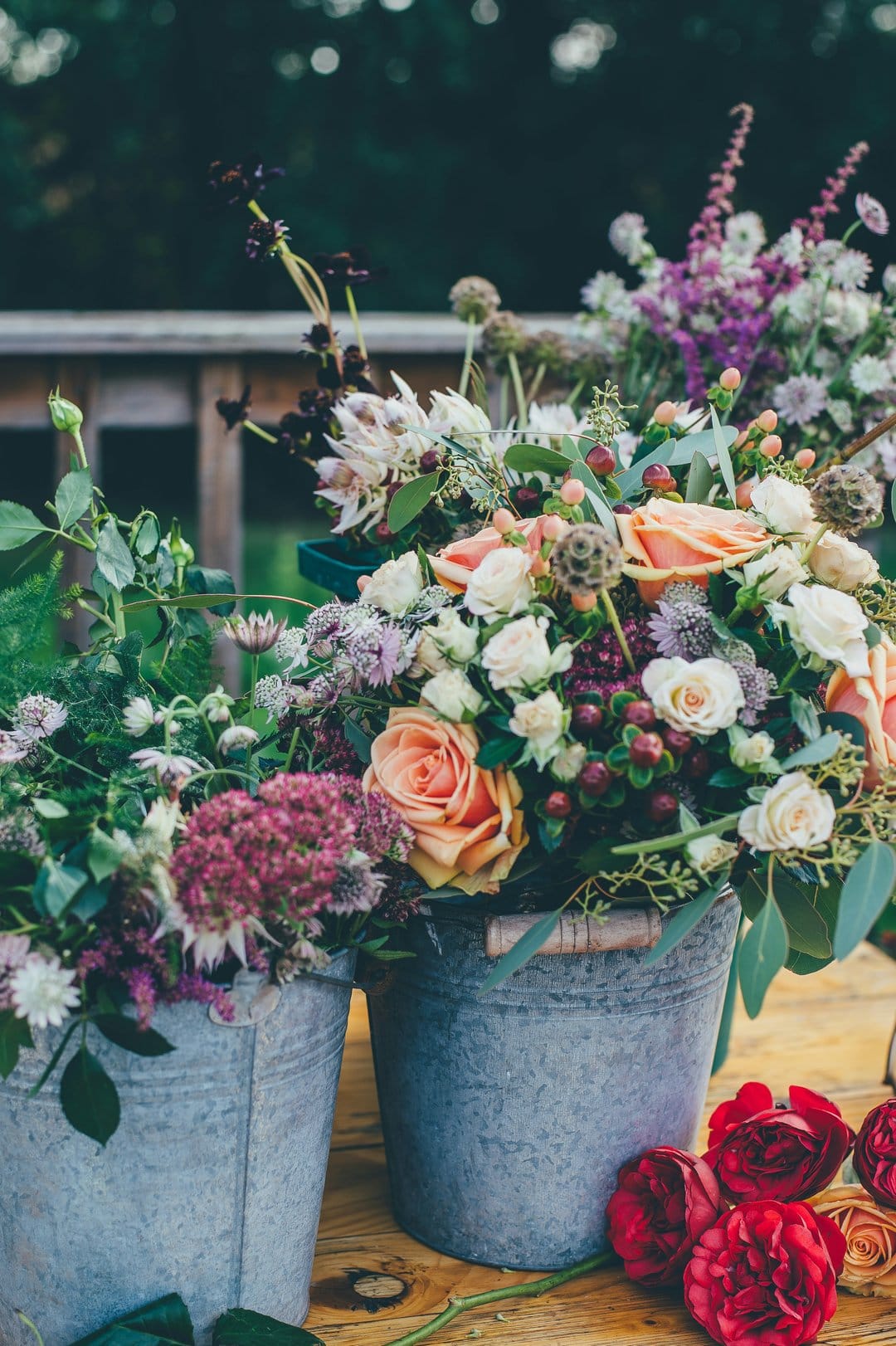 Two galvanized metal buckets filled with peach roses, pink sedum, and eucalyptus on a rustic wooden surface.