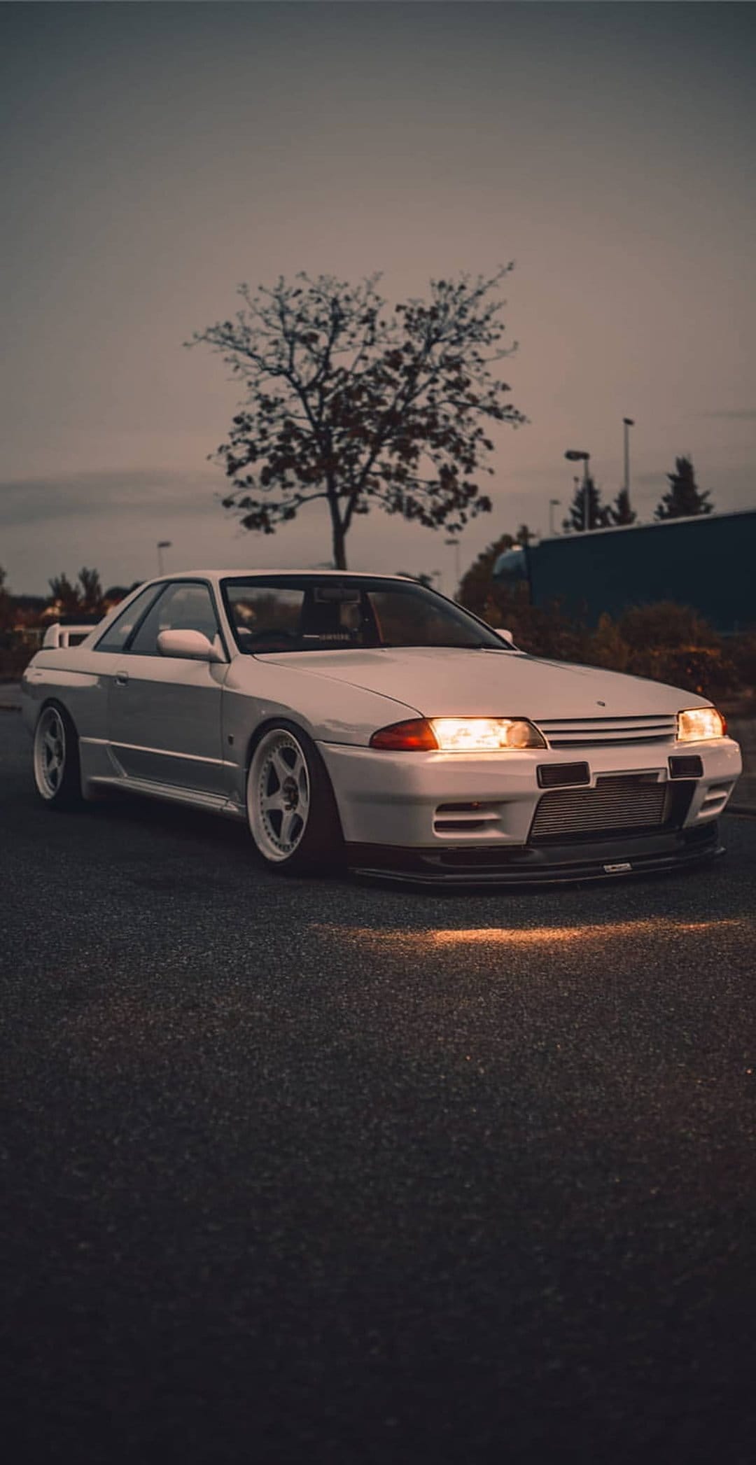 White Nissan Skyline R32 GTR with illuminated headlights parked on asphalt near a silhouetted tree at twilight.
