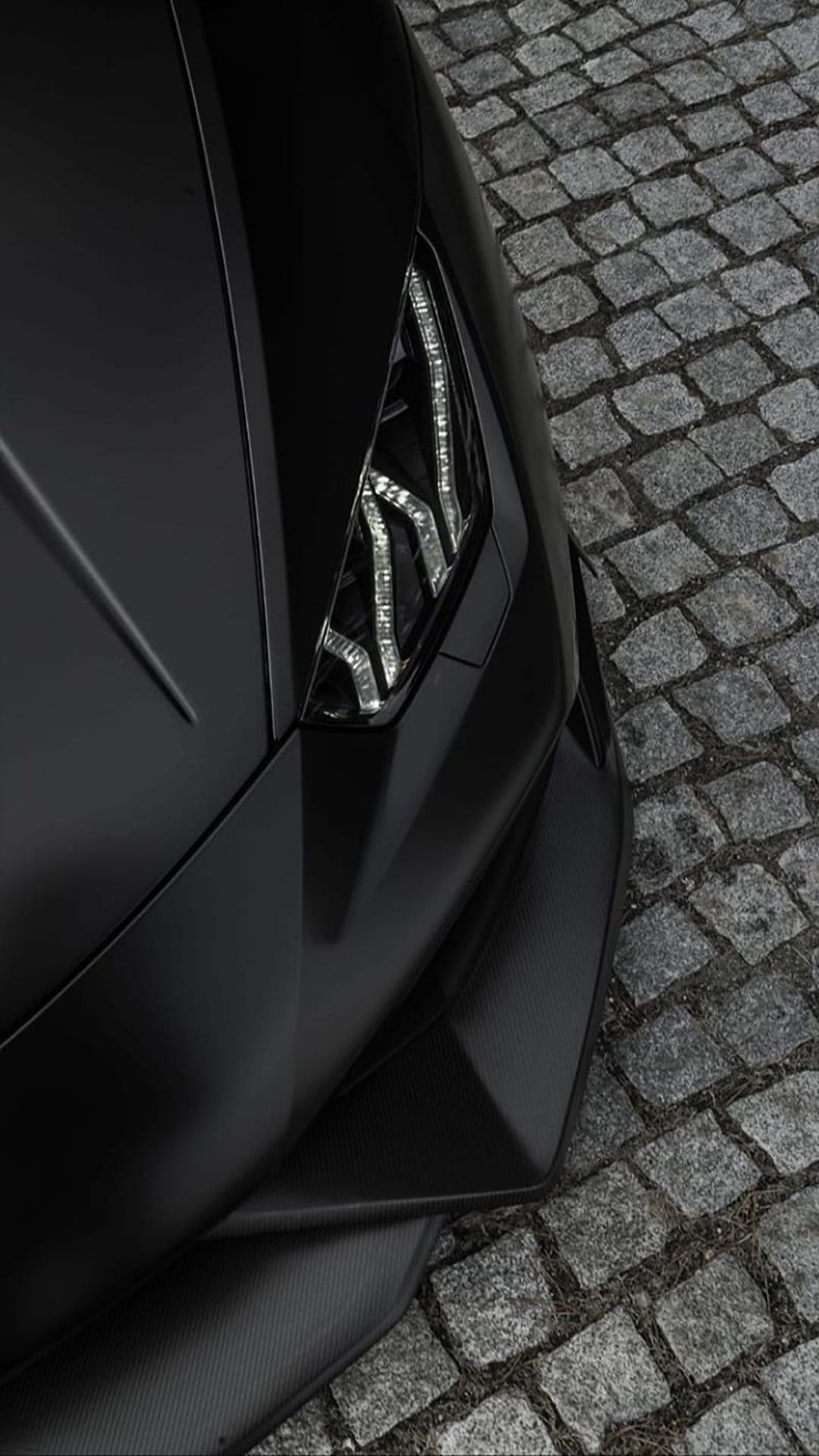 Matte black supercar headlight with glowing LED accents rests above a textured grey cobblestone road.