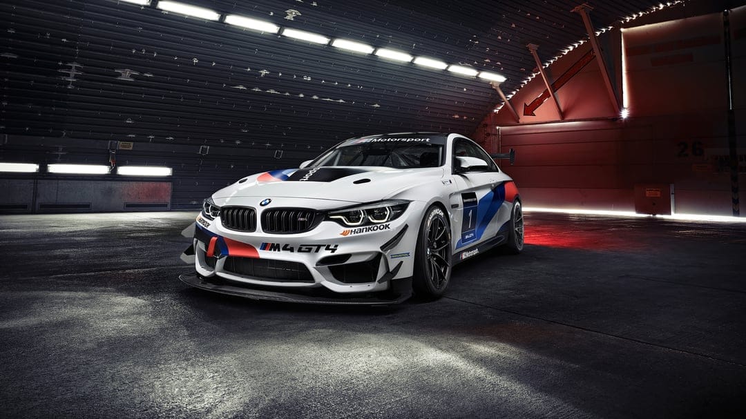 A white BMW M4 GT4 race car with M-Performance stripes in a dark industrial hangar under bright lights.
