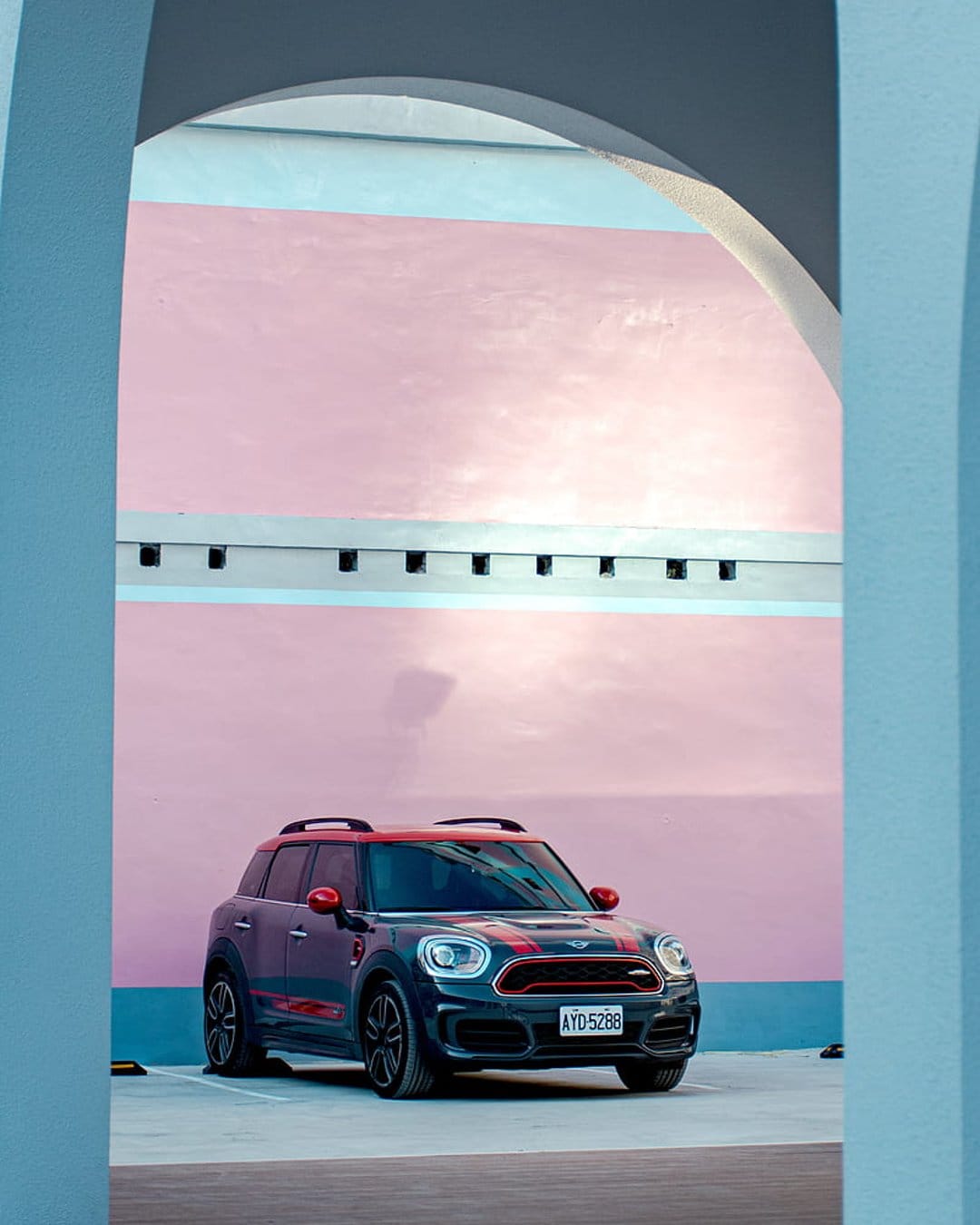 Black Mini Cooper Countryman with red racing stripes parked in front of a pink and blue striped textured wall.