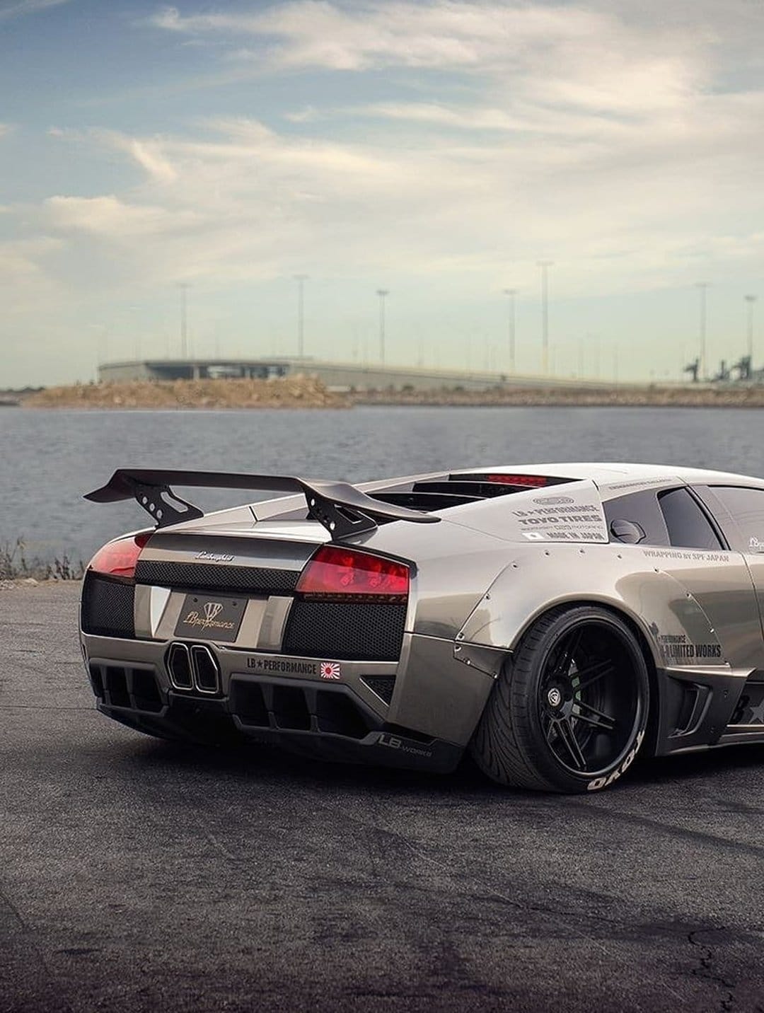 Lamborghini Murcielago with a widebody kit and large rear wing parked on tarmac near water under a cloudy sky.