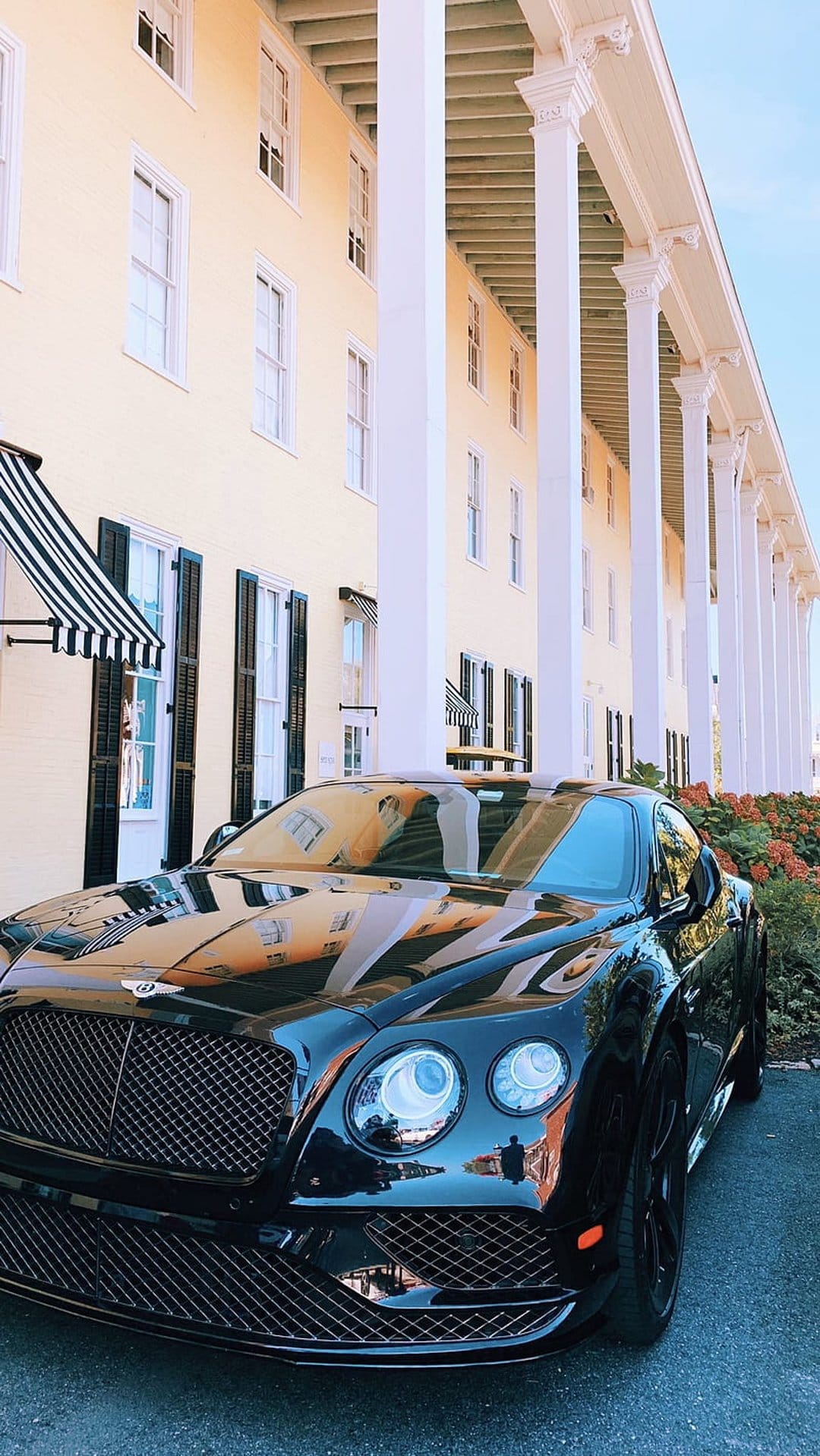Black Bentley Continental GT parked near a yellow neoclassical building with white columns and striped awnings.
