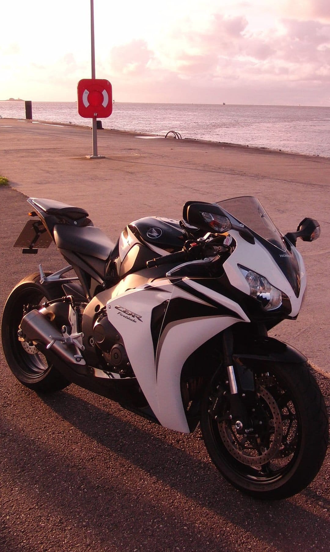 White and black Honda CBR motorcycle parked on a seaside walkway under a pink and orange sunset sky.