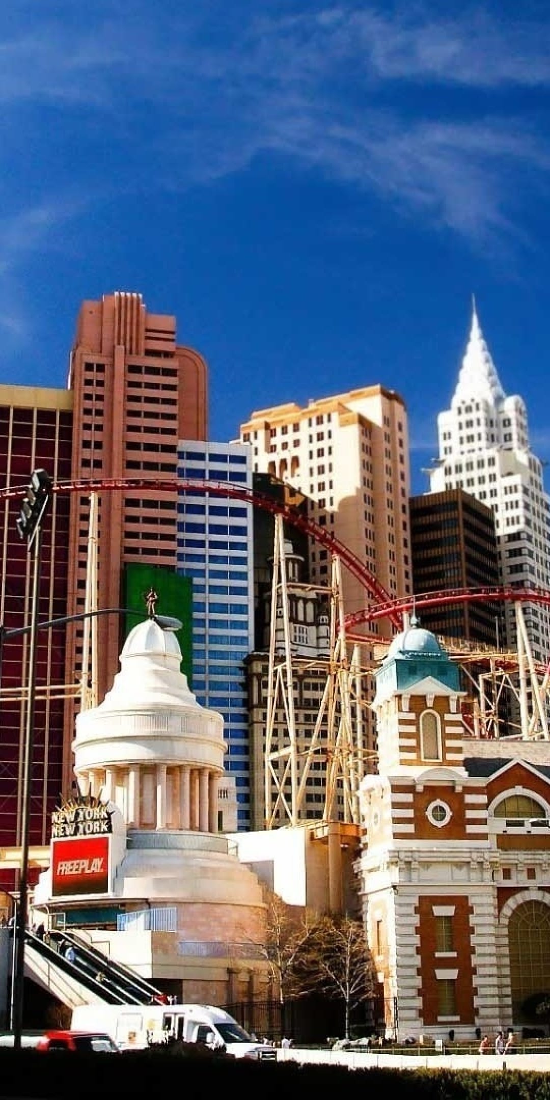 Red roller coaster tracks loop through architectural skyline replicas under a clear blue sky in Las Vegas.