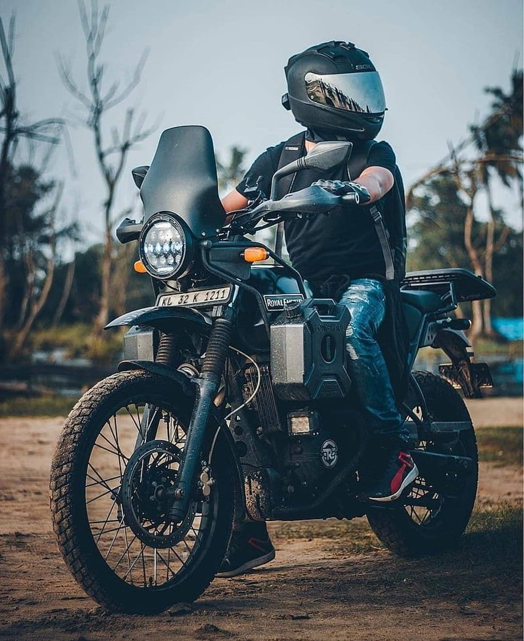 Biker on a black Royal Enfield Himalayan with auxiliary tanks on a dirt path under a hazy, overcast sky.
