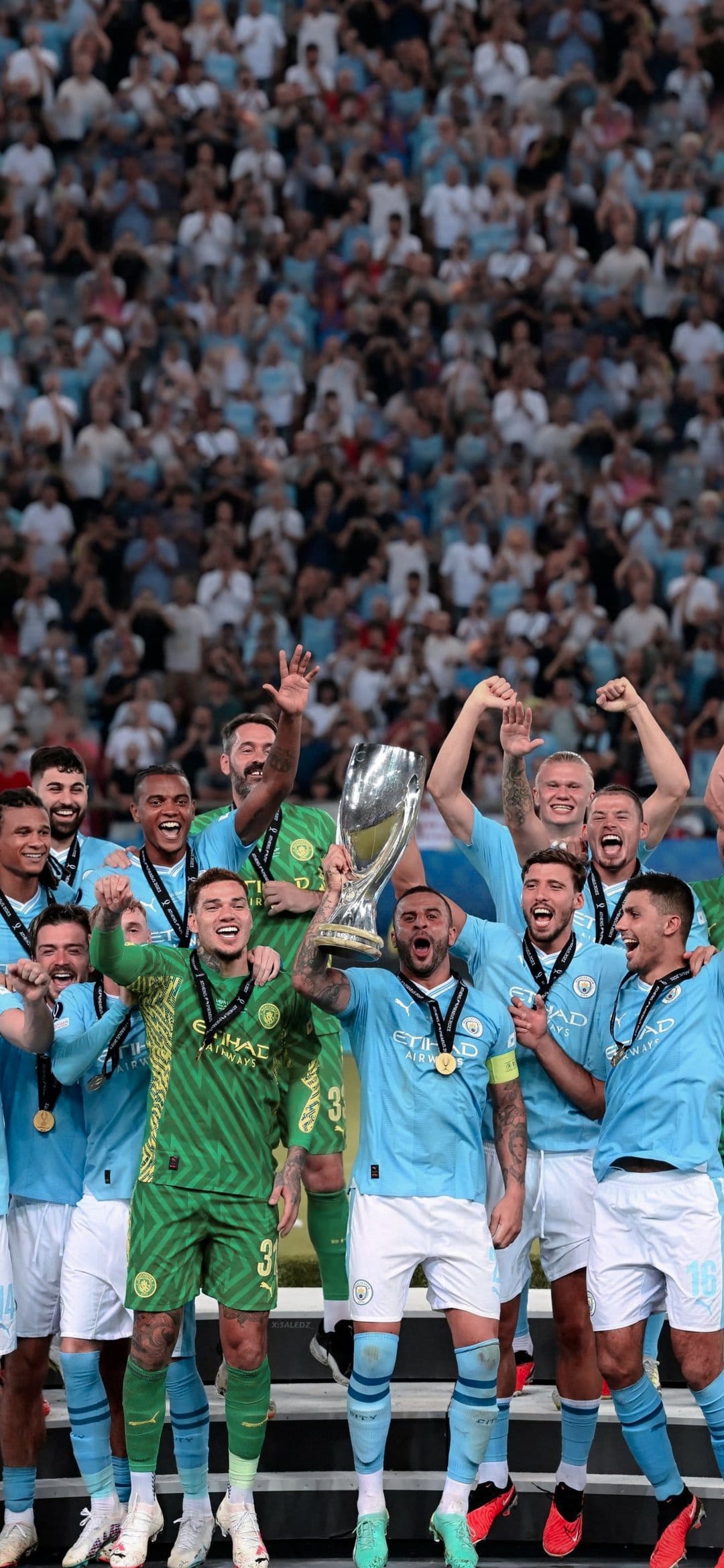 Manchester City players lift the silver UEFA Super Cup trophy overhead while cheering in a crowded stadium.