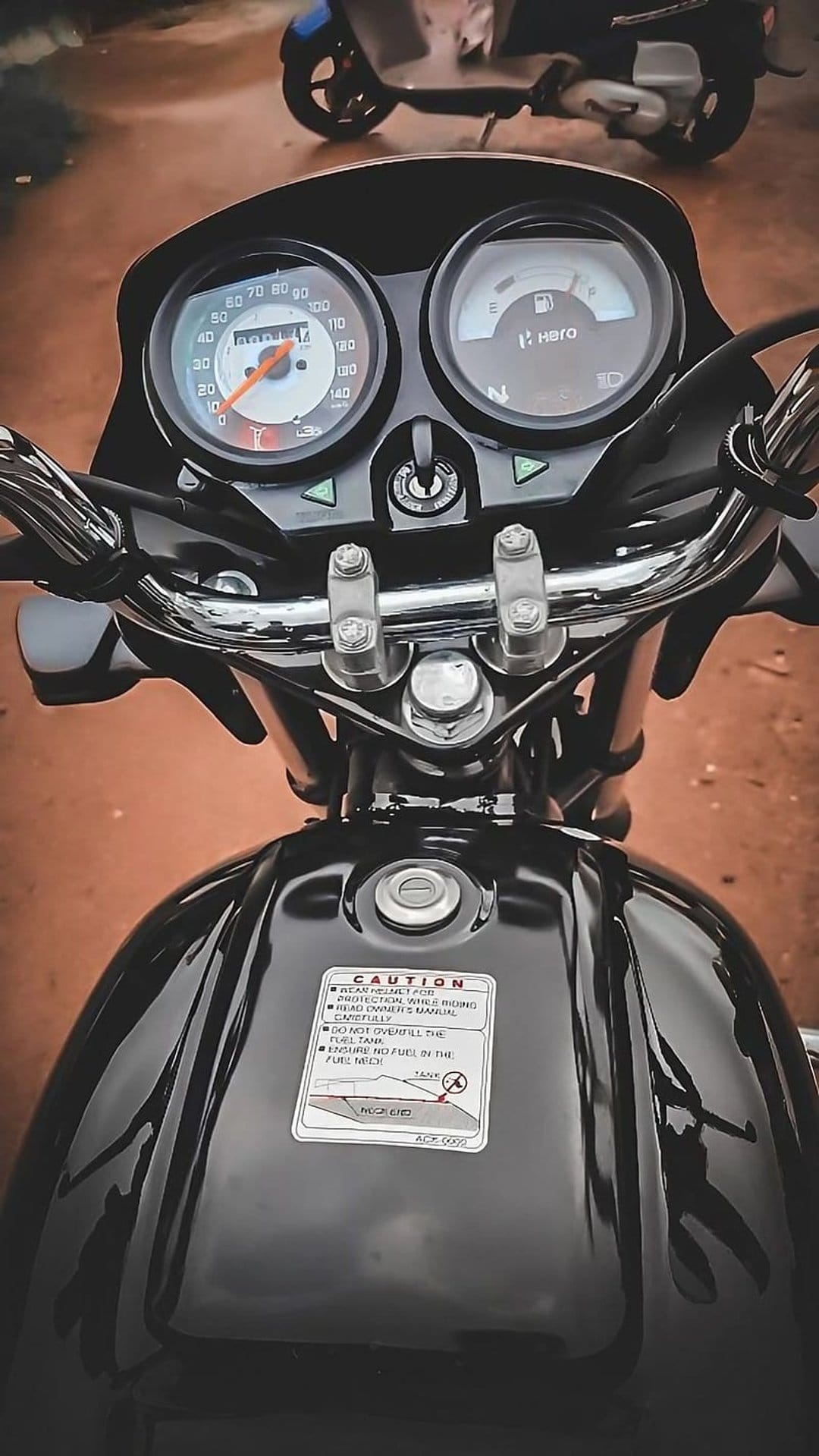 Black Hero Splendor motorcycle fuel tank and analog speedometer with a yellow caution sticker and chrome details.