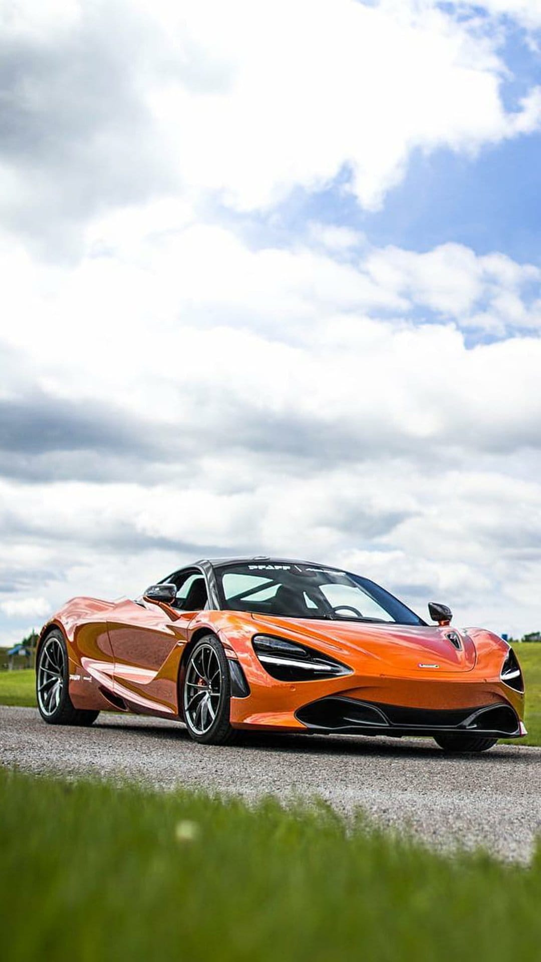 Orange McLaren 720S speedster parked on an asphalt road with green grass and a cloudy sky background.