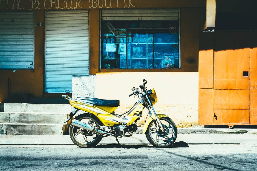 Yellow scooter with a black seat parked on asphalt in front of an orange building with blue-tinted windows.