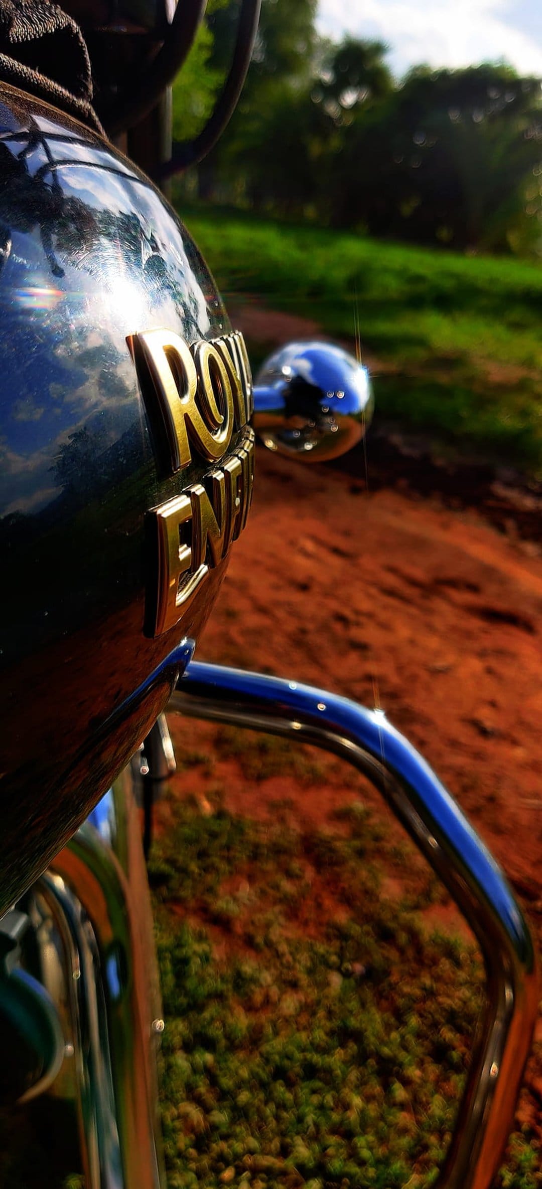 Royal Enfield motorcycle chrome handlebars and emblem reflecting green trees and blue sky on a dirt path.