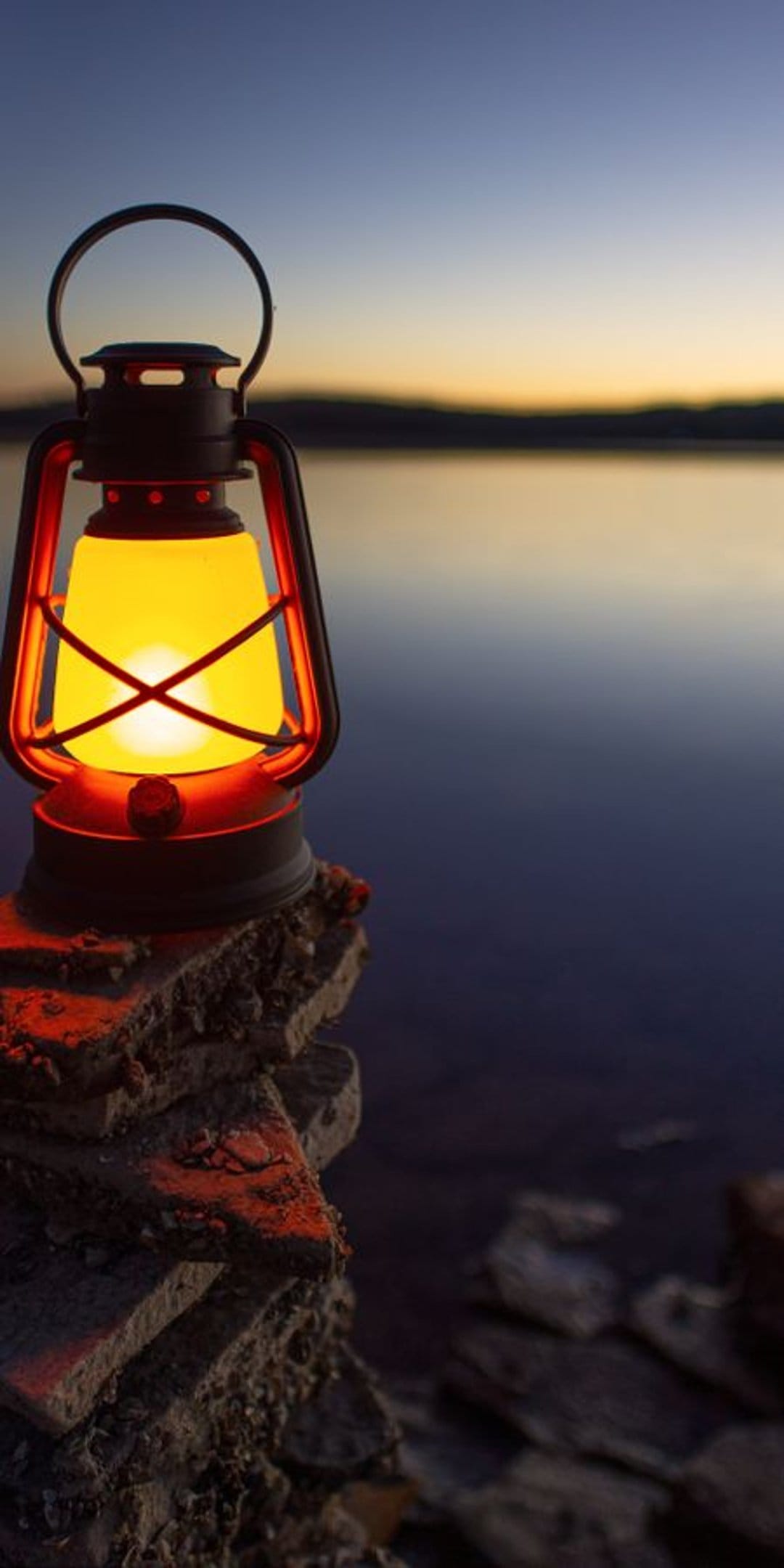 Orange lantern glows on stacked rocks beside a tranquil lake under a blue and violet evening sky.