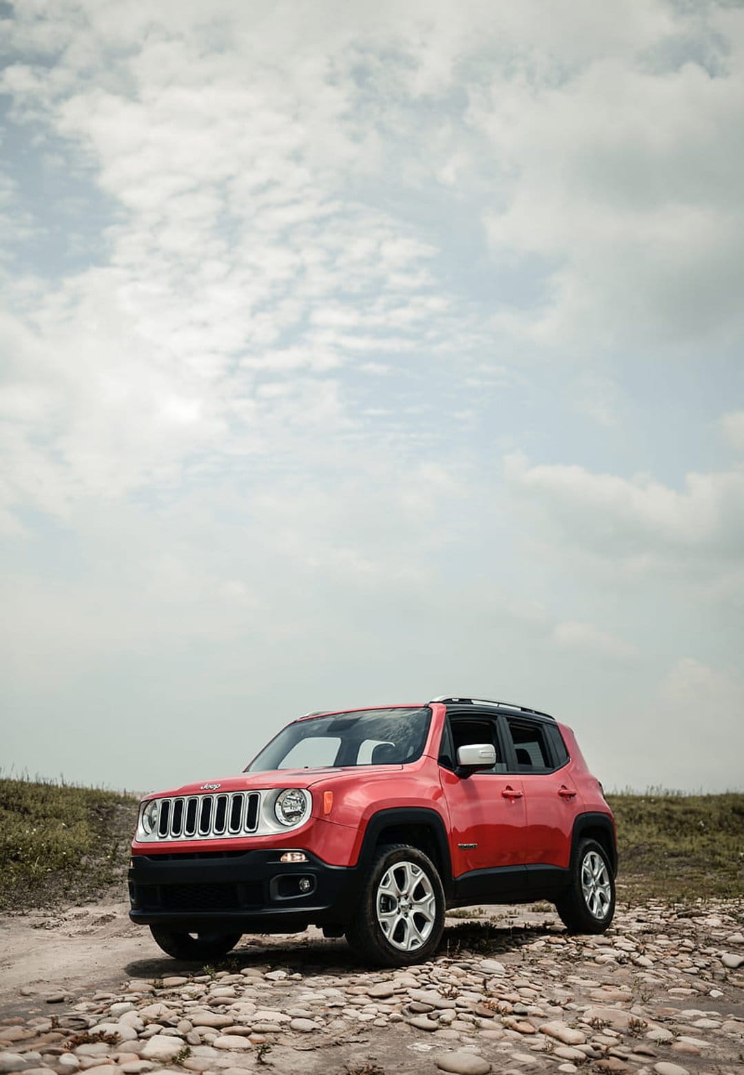 Red Jeep Renegade SUV parked on a gravel path under a cloudy sky with green grass visible in the background.