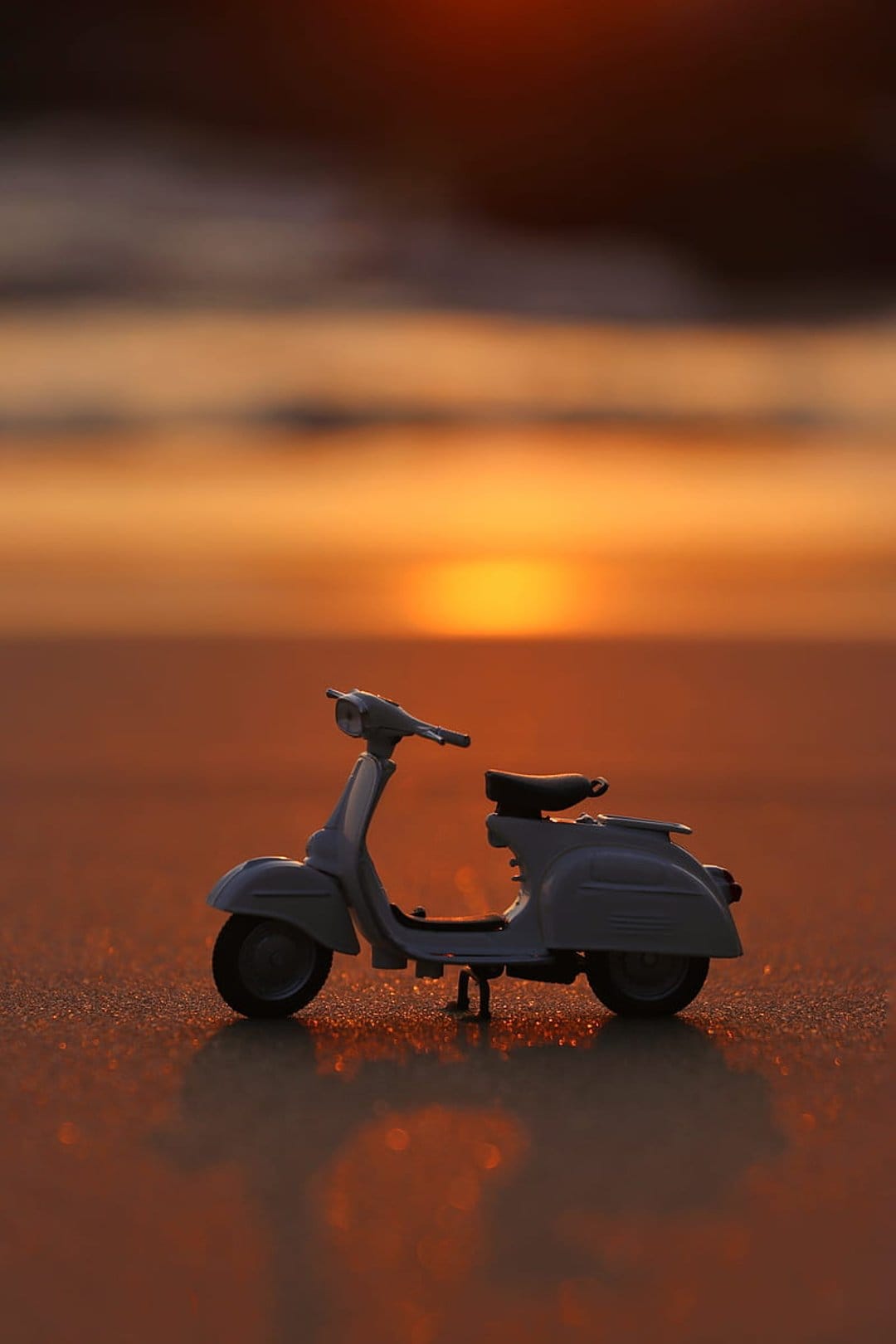 Miniature blue Vespa scooter resting on wet beach sand reflects the orange and pink sunset sky during dusk.
