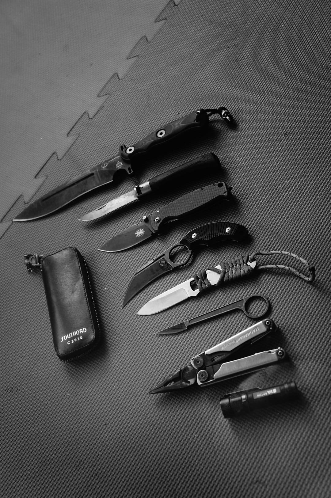 Folding knives, a multi-tool, and a flashlight rest on a dark textured surface in a monochrome top-down view.