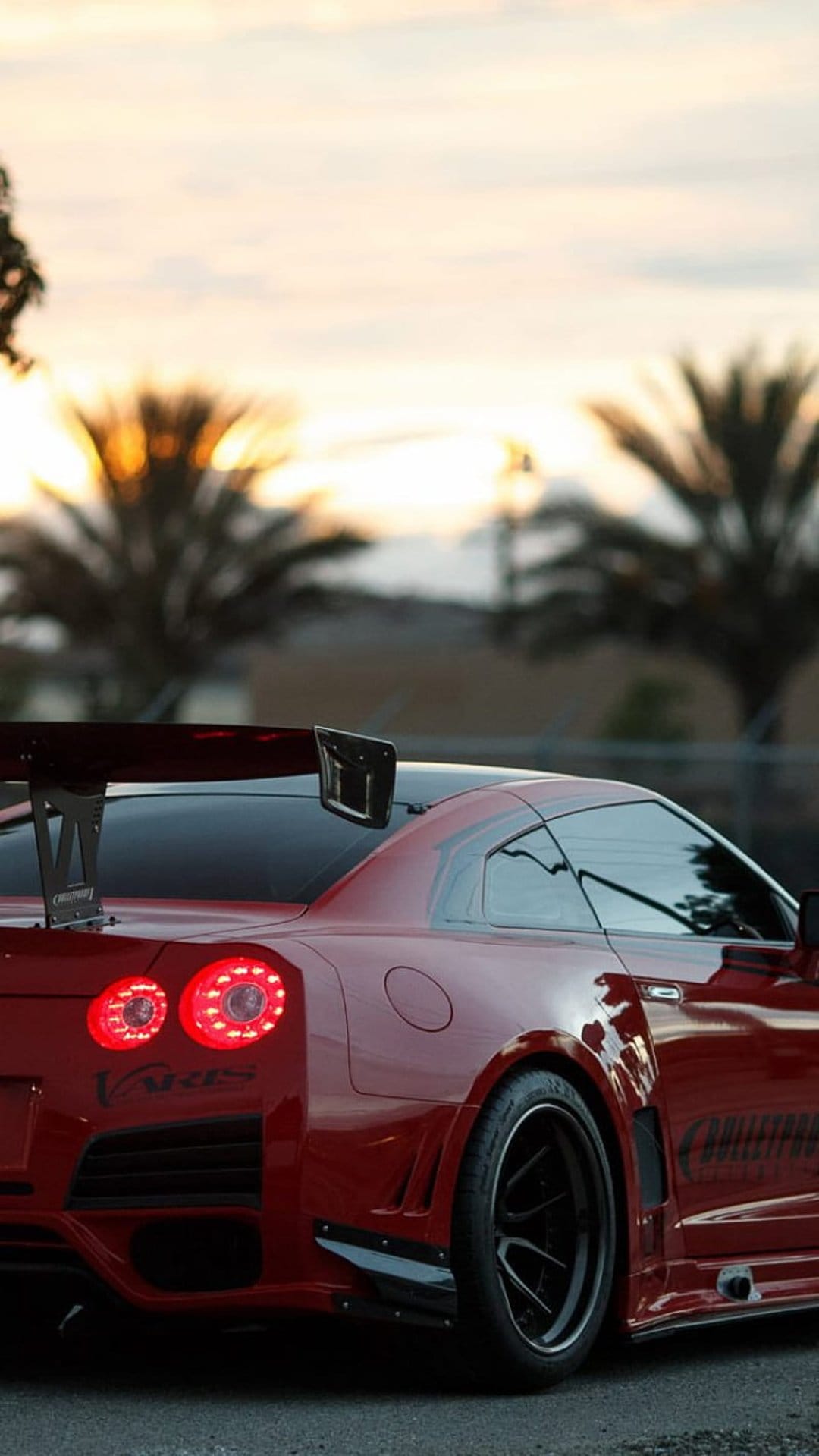 Red Nissan GT-R sports car with a large rear wing parked under palm trees during a warm golden hour sunset.
