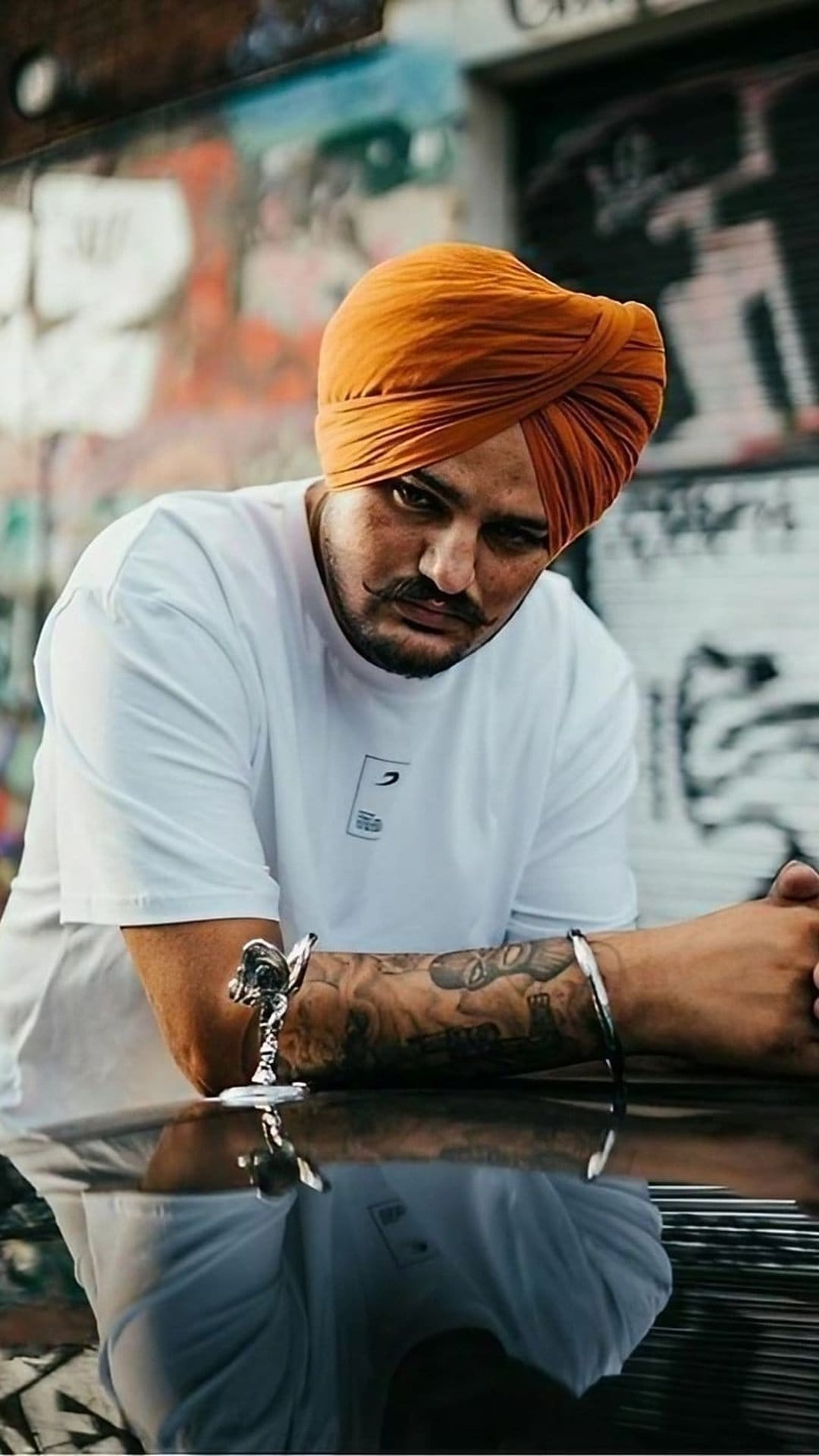Sidhu Moose Wala wears an orange turban and white t-shirt with detailed arm tattoos and a silver bracelet.