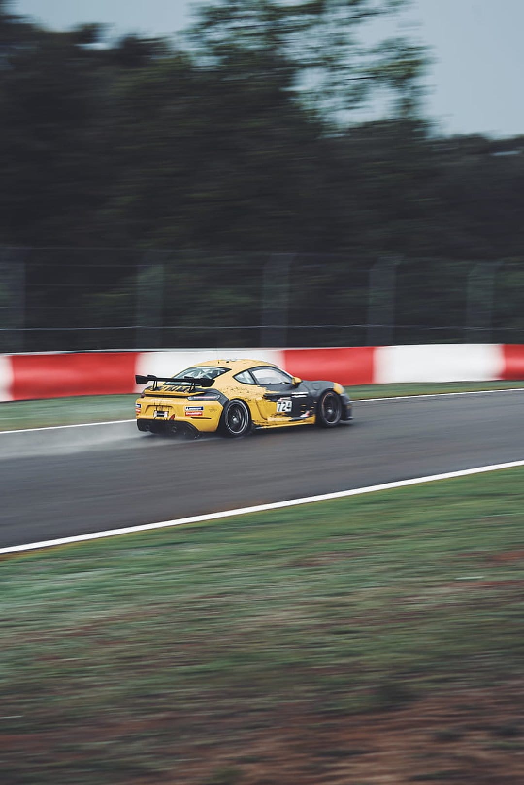 Yellow Porsche race car accelerates on a wet circuit with motion blur against red and white trackside barriers.
