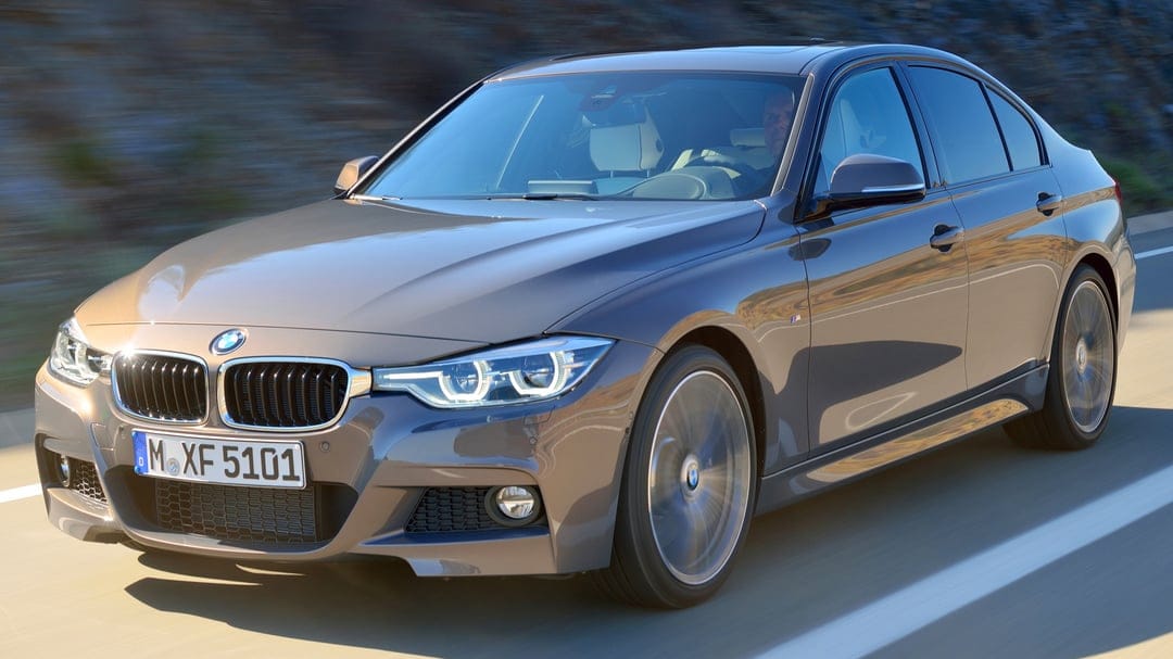 Bronze BMW 3 Series sedan driving fast on a sunlit road with heavy motion blur on wheels and background.