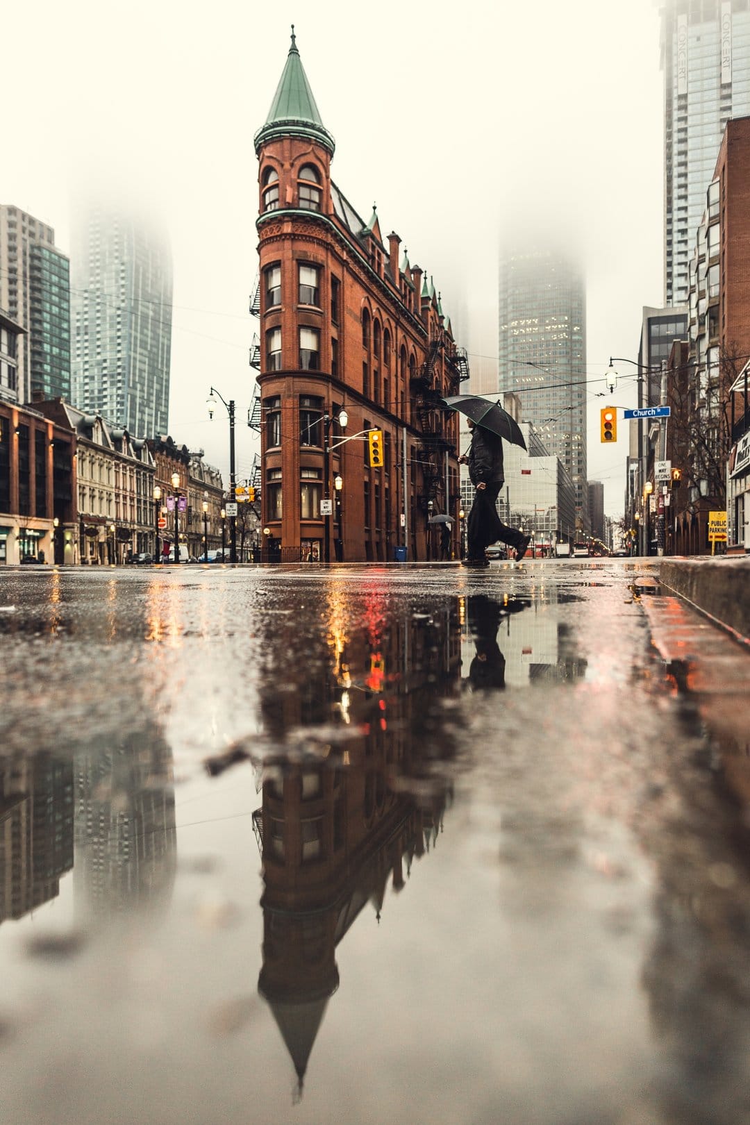 Lone figure with a dark umbrella crosses a wet city street near the Flatiron Building during a foggy evening.