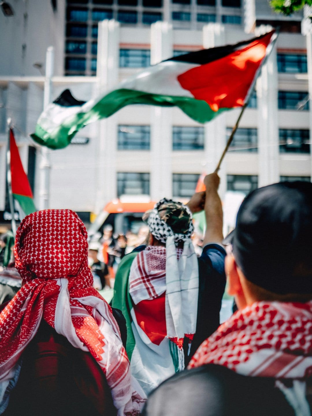 Protesters from behind wave Palestinian flags in a city street near a bus and buildings during a public rally.
