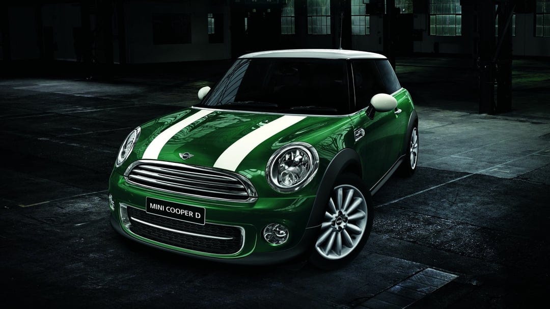 Green Mini Cooper D with white racing stripes parked in a dark industrial warehouse with reflective floors.