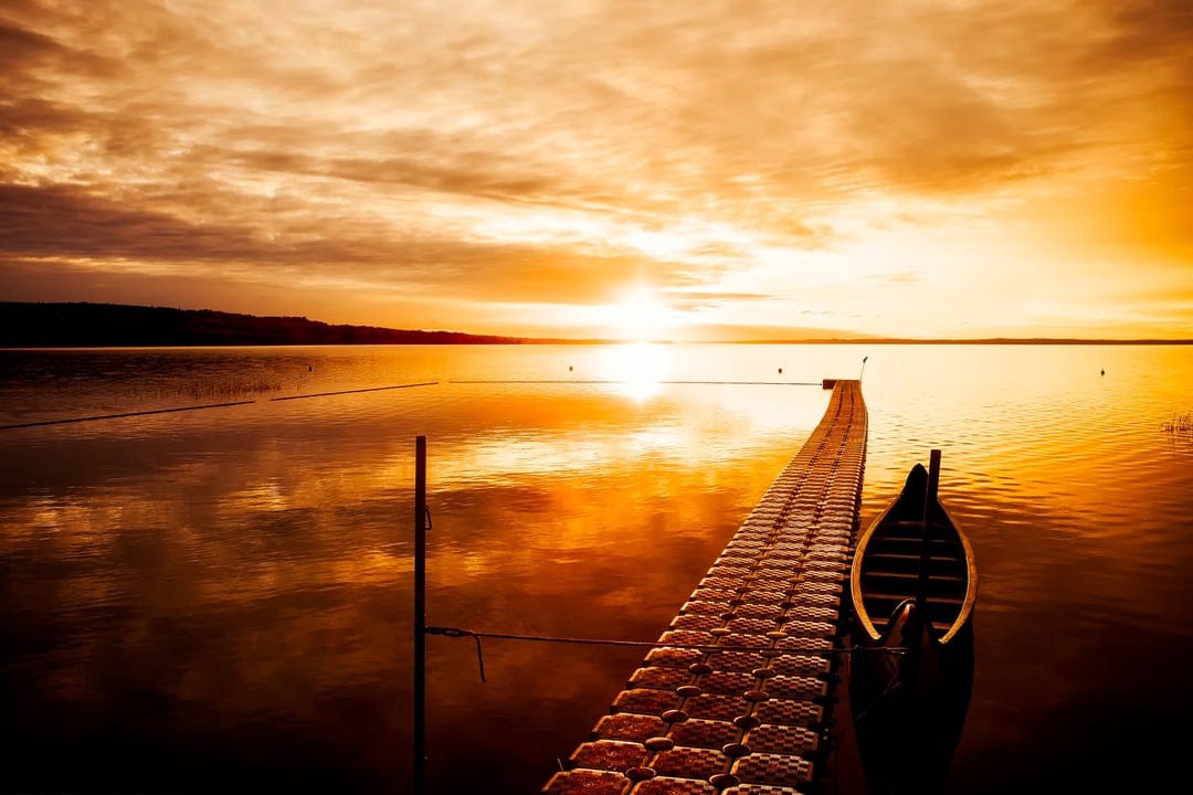 Wooden pier and small boat on a still lake reflecting a dramatic orange sunset with heavy atmospheric clouds.