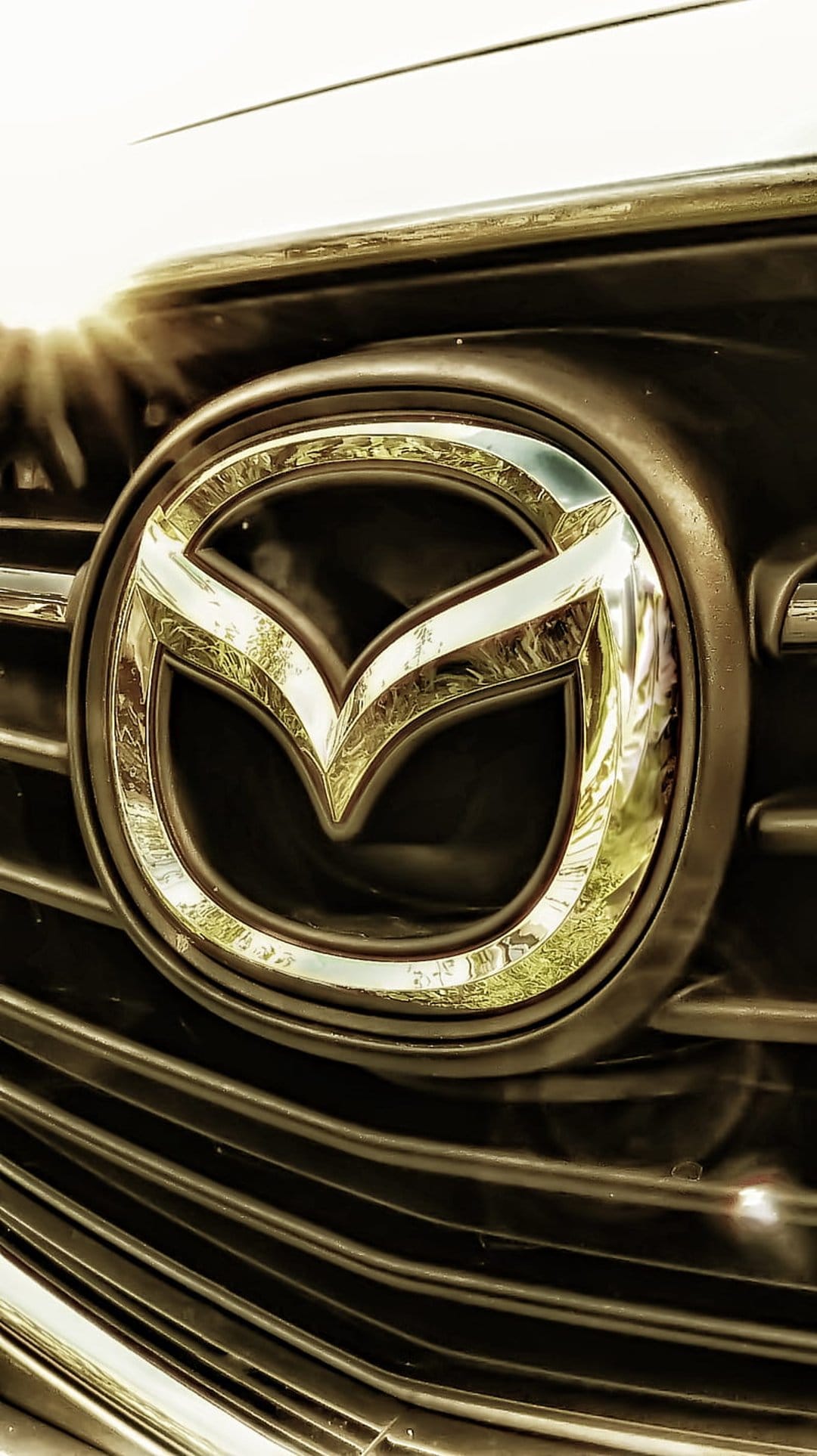 Mazda chrome logo emblem on a black mesh grille with a bright white sun flare and metallic reflections.