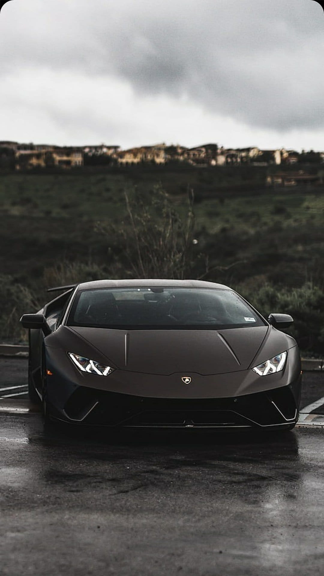 Dark Lamborghini supercar parked on wet asphalt with glowing headlights against a cloudy hill landscape.