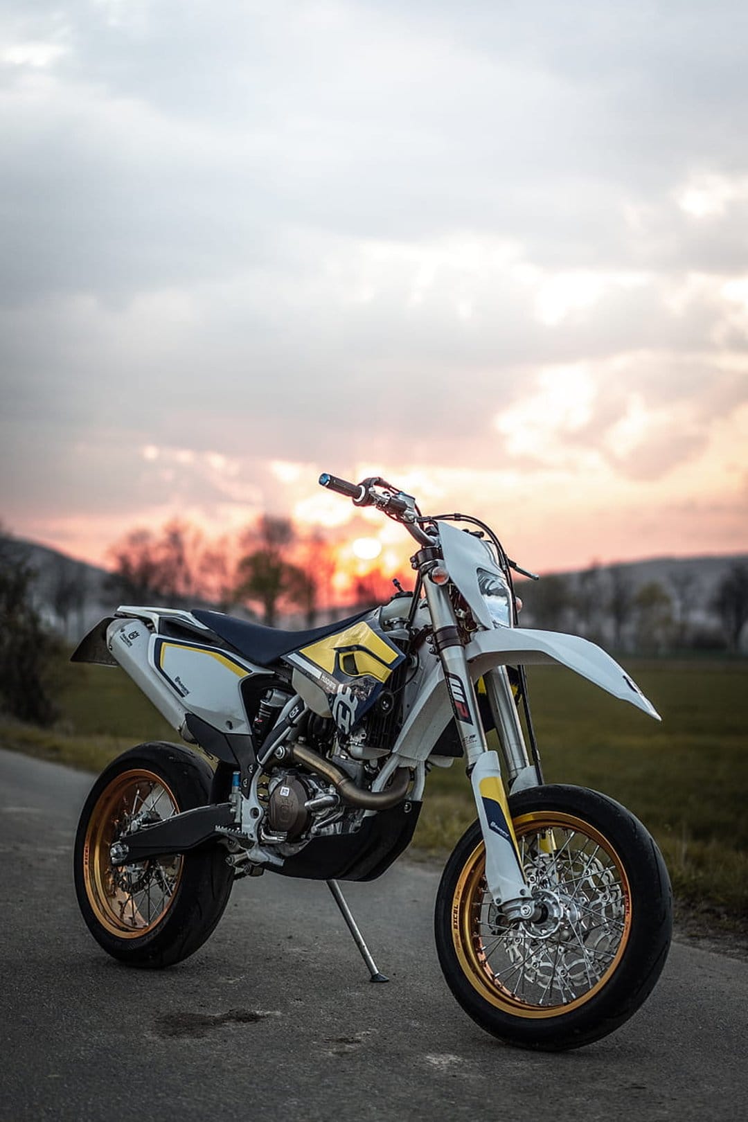 Supermoto motorcycle with golden rims parked on a paved road during a vibrant sunset with soft colorful clouds.