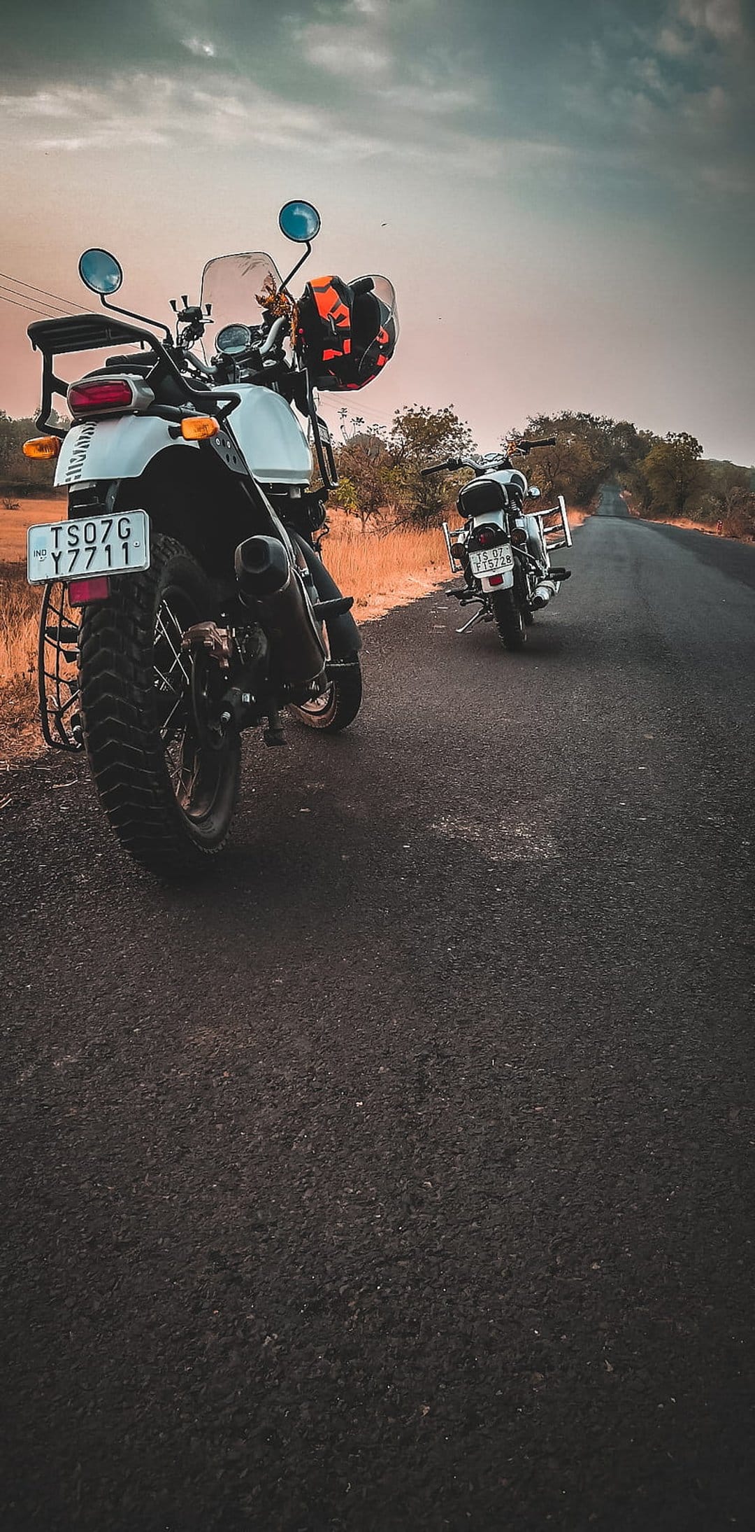 Two Royal Enfield motorcycles with knobby tires parked on a rural road during a dramatic golden hour sunset.