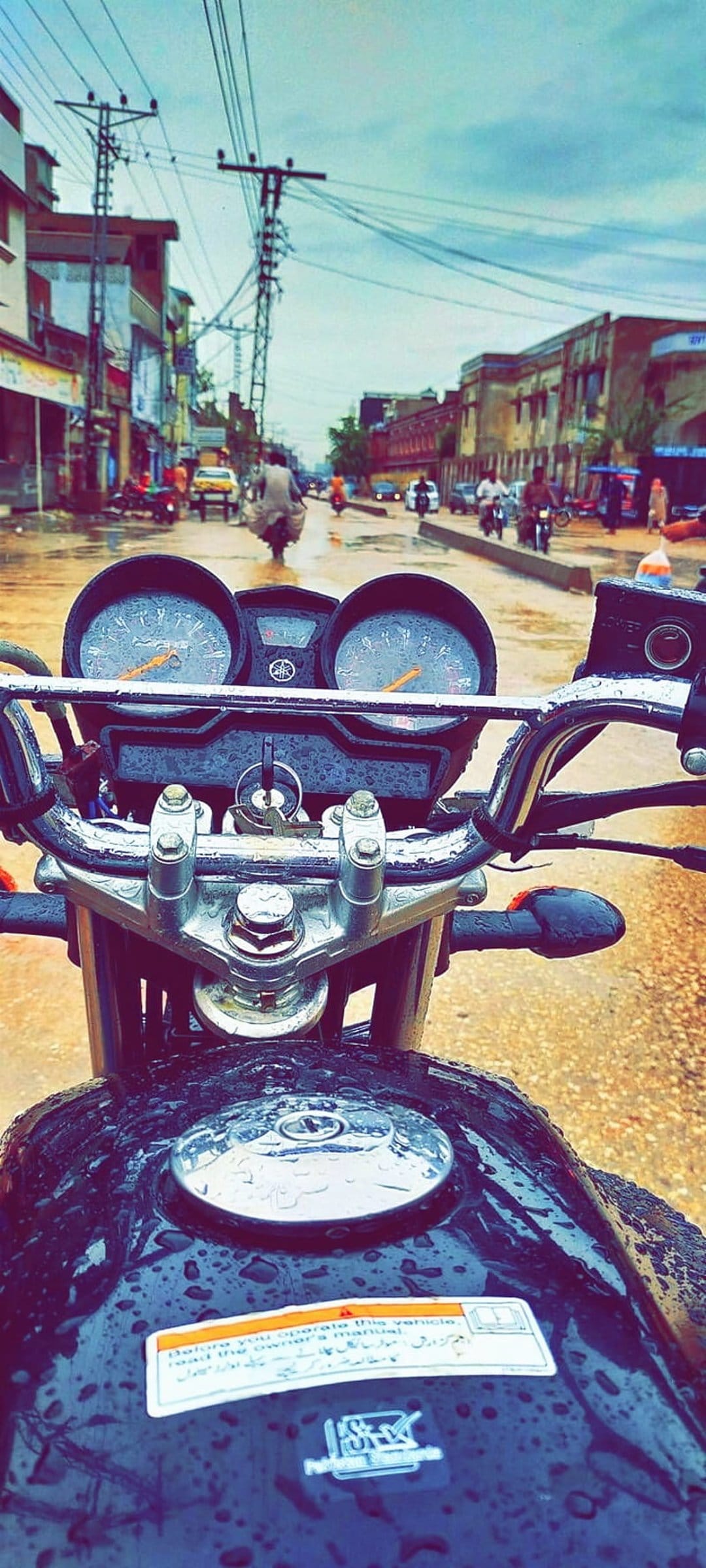 Motorcycle fuel tank and dashboard covered in raindrops on a wet street with town buildings in the background.