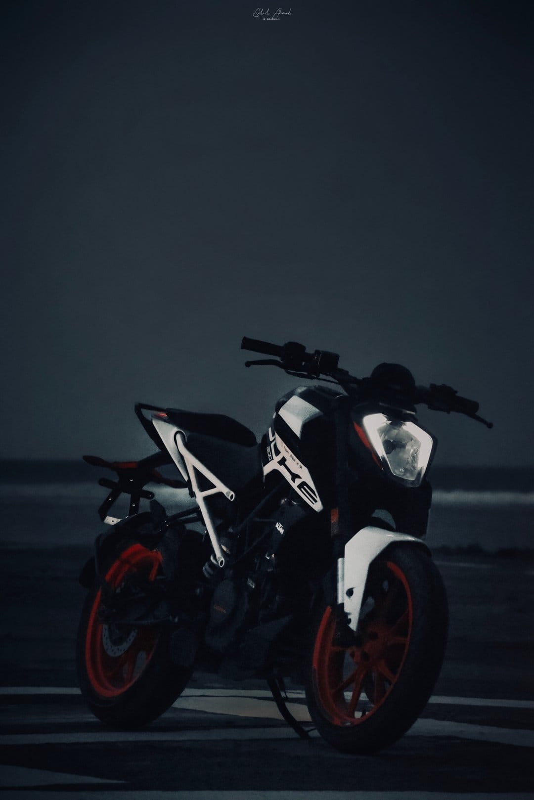KTM Duke motorcycle with orange wheels parked on a dark sandy beach under a moody overcast sky at twilight.