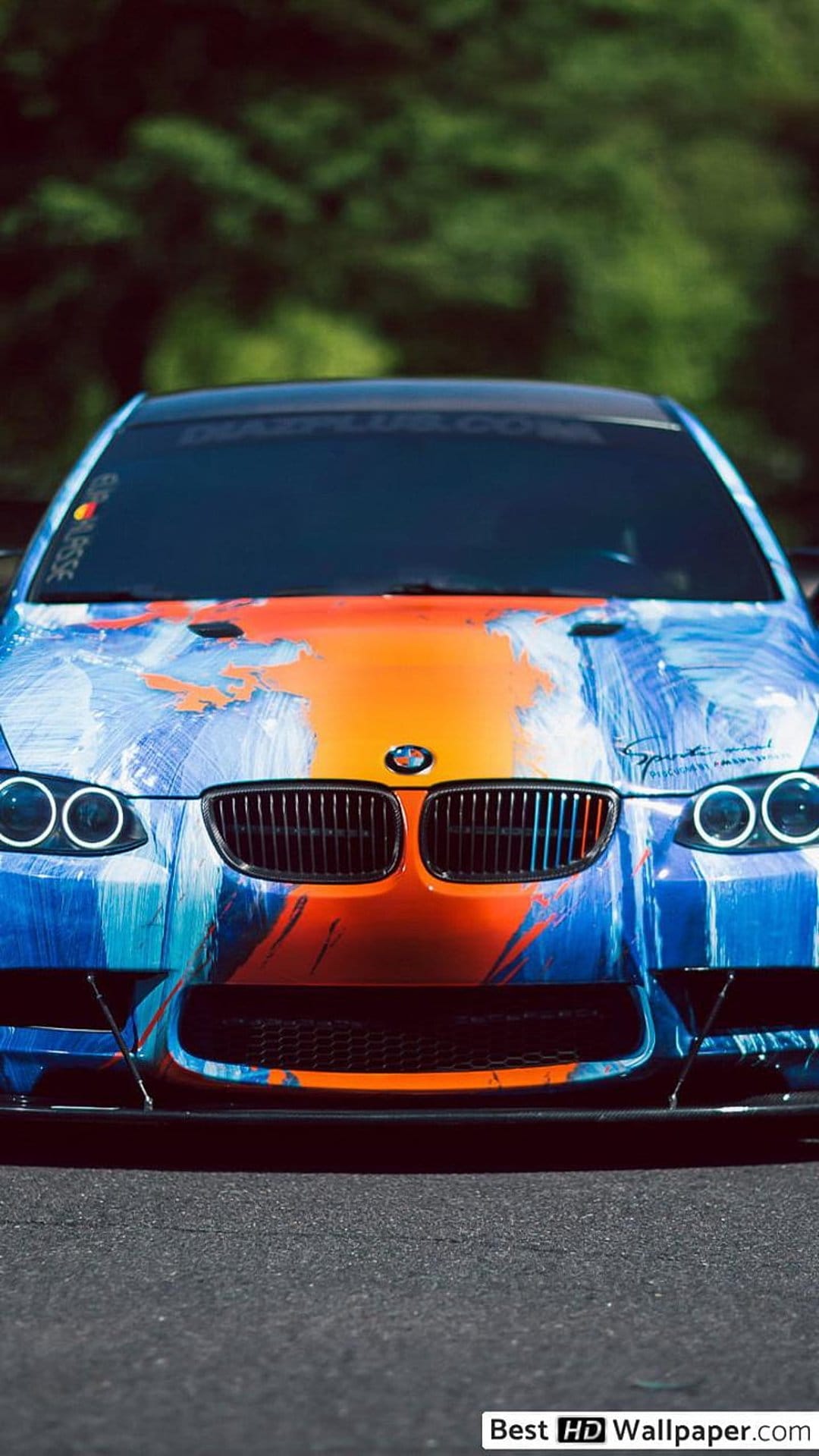 BMW M3 front view with blue and orange abstract paint splatters on an asphalt road with glowing headlights.