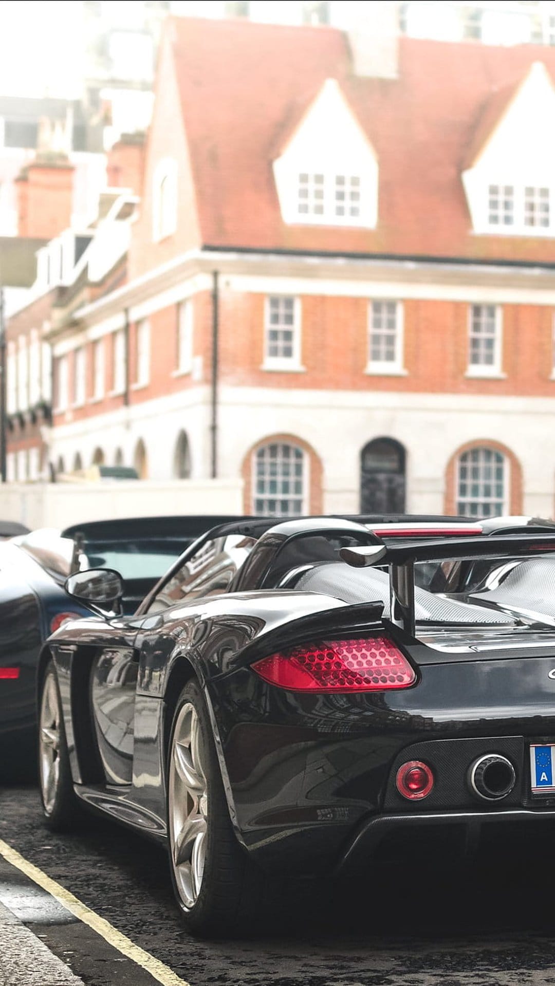 Black Porsche convertible with glowing red taillights parked on a European cobblestone street near brick buildings.