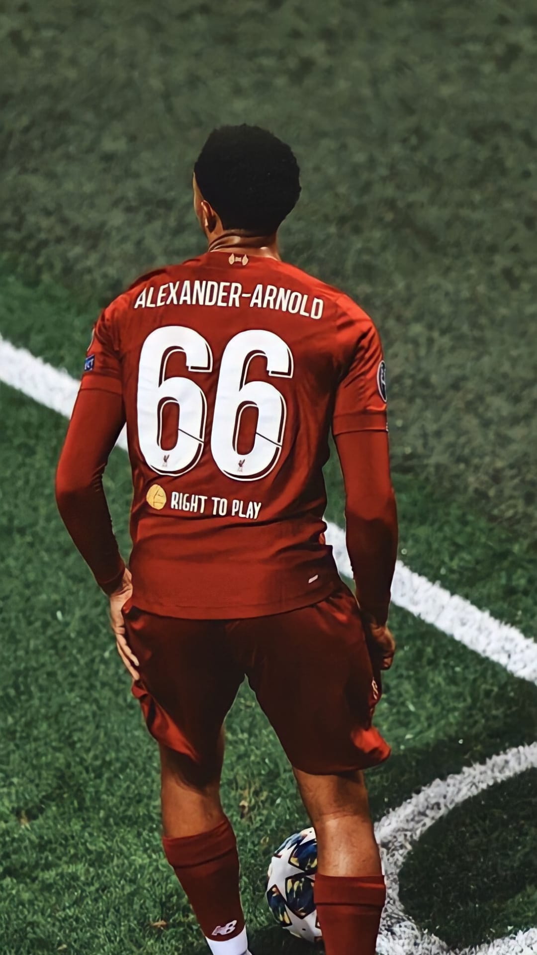 Trent Alexander-Arnold in a red 66 Liverpool jersey stands on a grass pitch with a football near his white cleats.