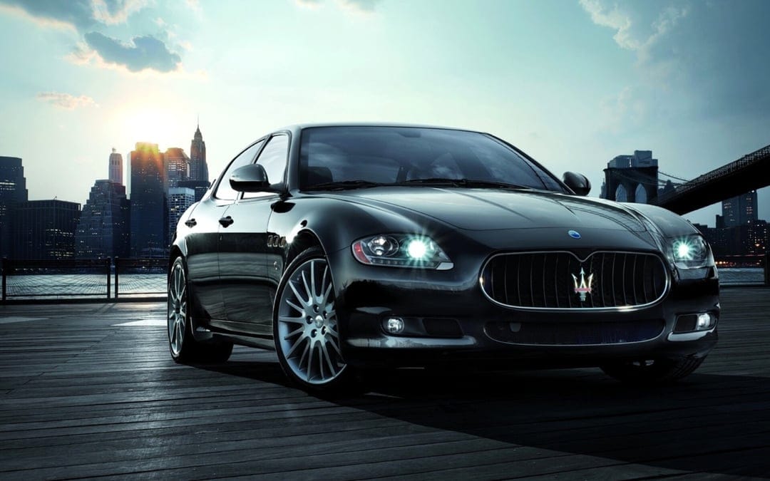 Black Maserati sedan with lit headlights parked on a wooden pier before a sunset city skyline and bridge.