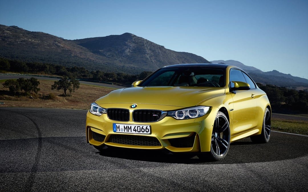 Yellow BMW M4 sports car parked on a paved road with rugged mountains and a clear blue sky background.