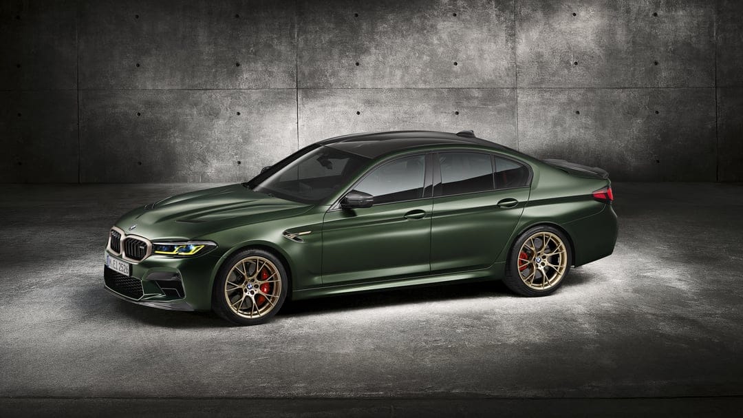 Dark green BMW M5 CS with gold wheels and red brake calipers against a grey concrete wall background.